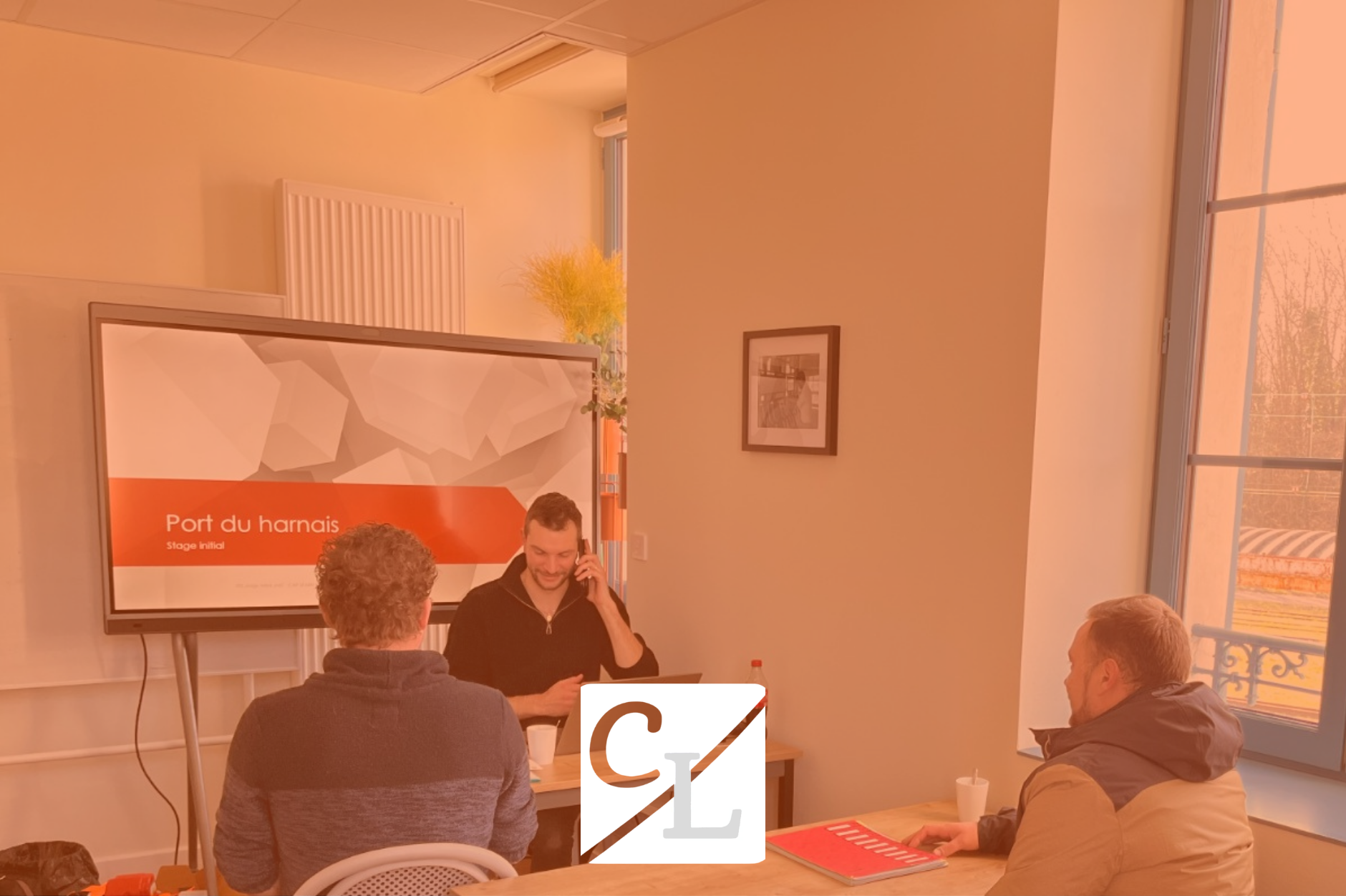 Une réunion se déroule dans une salle avec plusieurs personnes. Un homme parle au téléphone devant un écran affichant un panneau intitulé 'Port du harnais', avec d'autres personnes assises autour de la table.