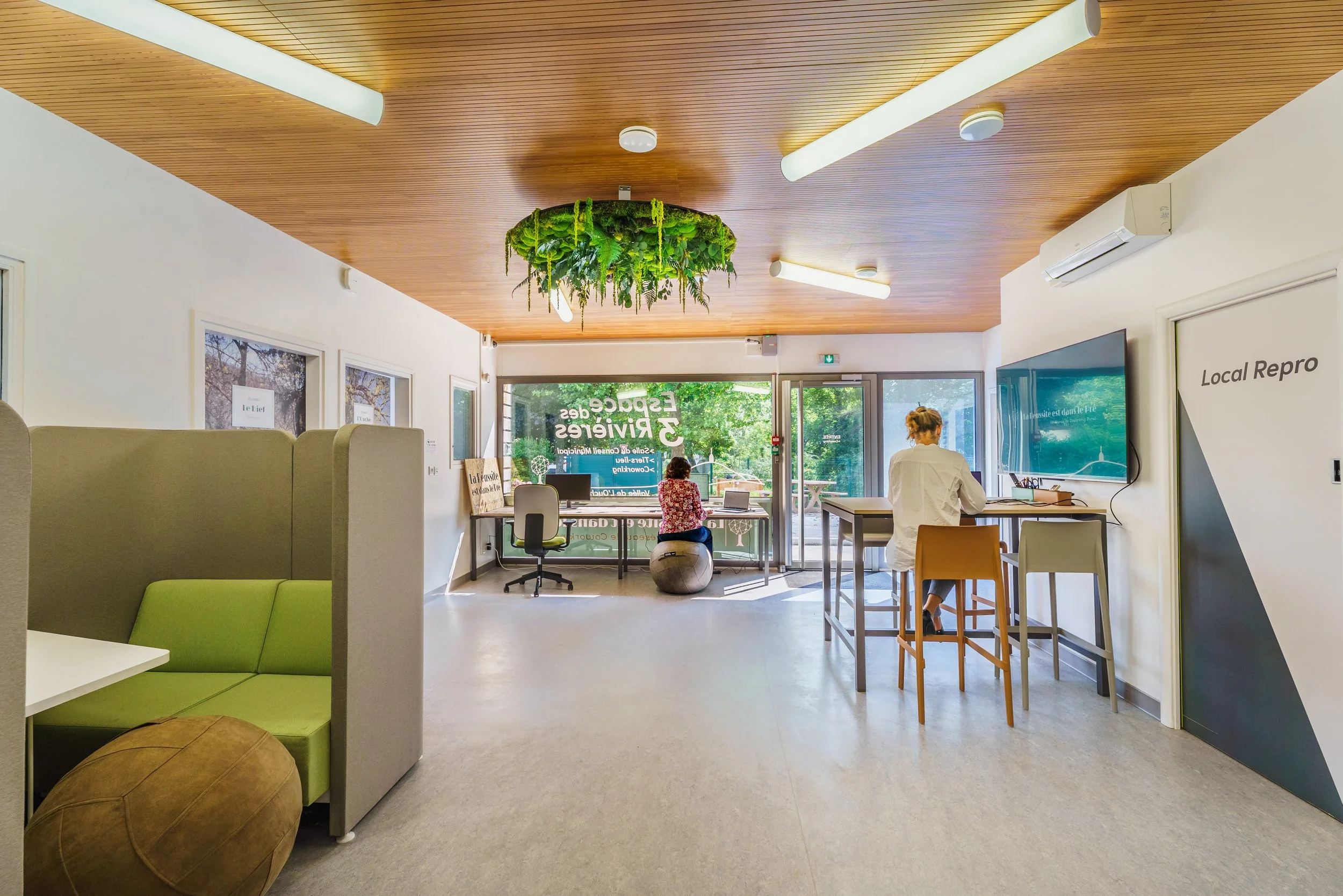 Intérieur d'un espace de bureau lumineux, avec un plafond en bois, un mur blanc, plusieurs personnes assises ou debout, un grand écran mural, un mur avec fenêtres, et une décoration végétale suspendue au plafond.