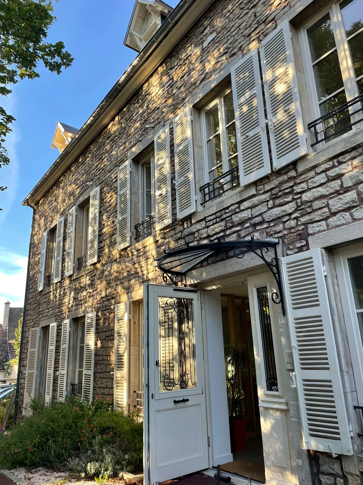 Maison en pierre avec fenêtres à volets blancs et ouverture de porte en métal noir, jardin avec plantes et fleurs.