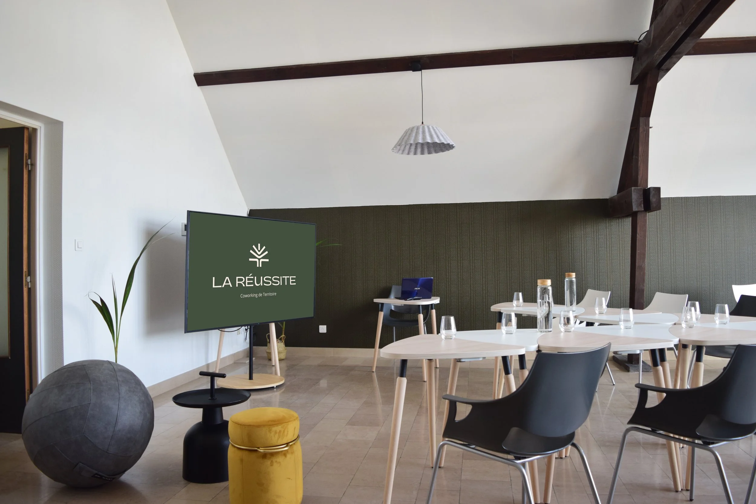 Salle de réunion avec tables rondes, chaises, écran affiche 'LA RÉUSSITE Coworking de Territoire', décoration moderne, plafond avec poutres en bois, lampe suspendue, plantes et bouteilles d'eau sur les tables.