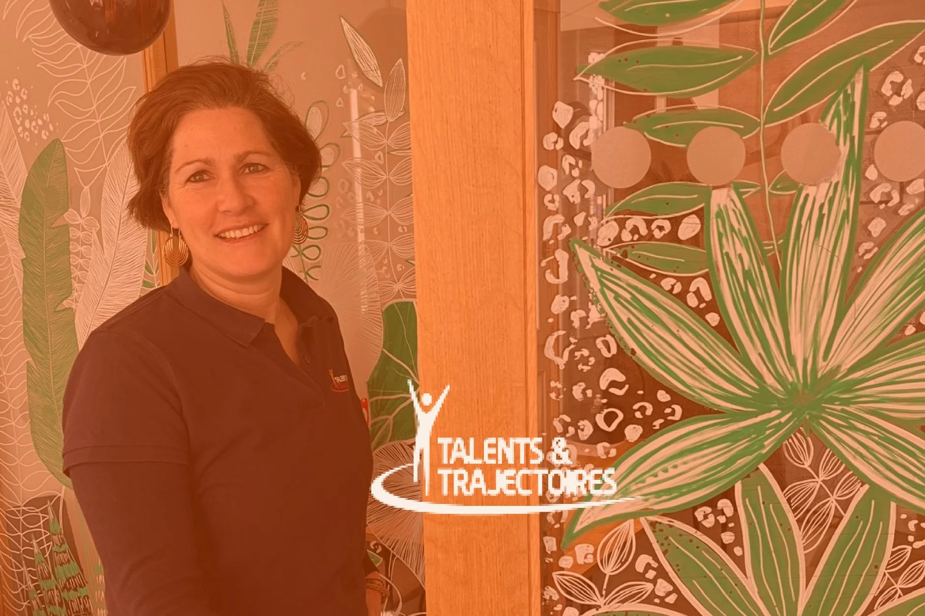 Une femme souriante portant un chemisier noir se tient devant un mur décoré de motifs végétaux et une affiche avec le texte 'Talents & Trajectoires'.