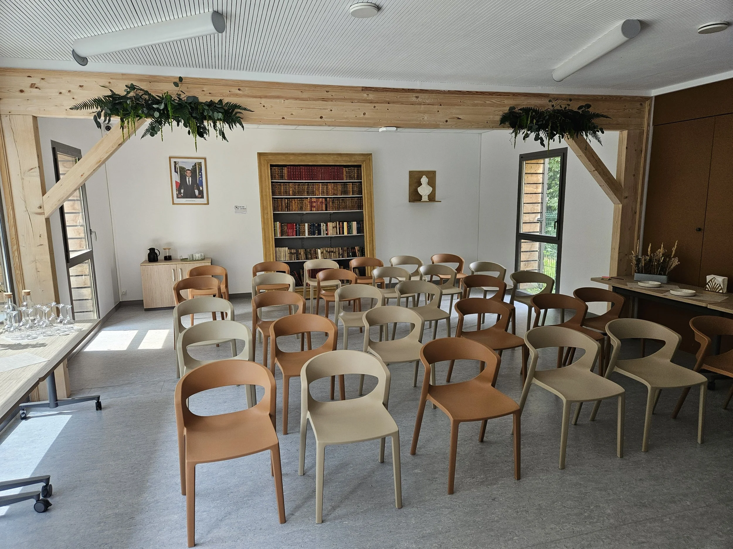 Salle de réunion ou salle d'attente avec plusieurs chaises en bois beige et marron, bibliothèque pleine de livres, tableaux, et décorations murales, fenêtres laissant entrer la lumière naturelle, style moderne et chaleureux.