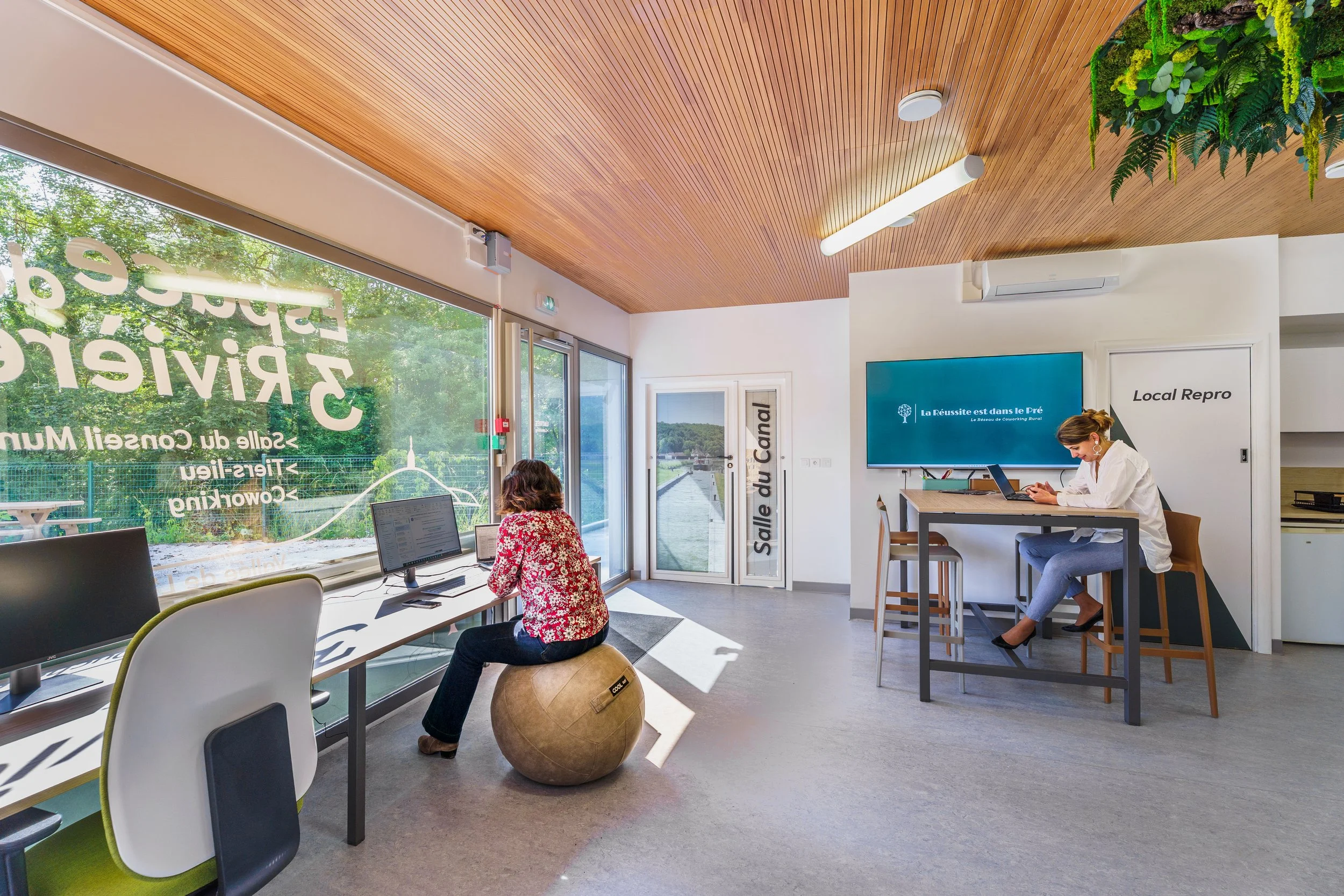 Intérieur d'un espace de coworking avec deux femmes travaillant sur des ordinateurs, beaucoup de lumière naturelle, murs blancs, plafonds en bois, et une végétation extérieure visible par la grande vitre