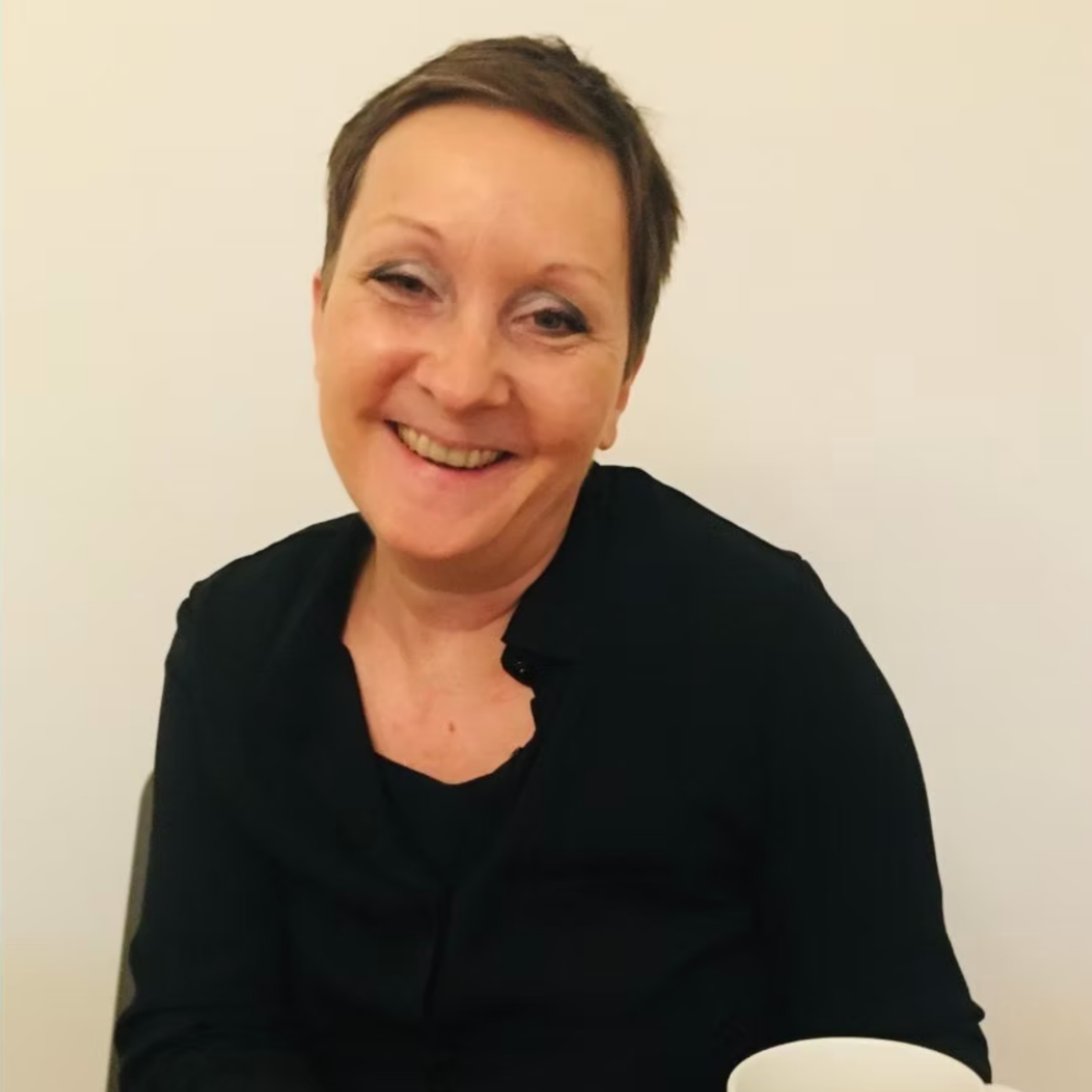 Femme souriante à courte chevelure brune, portant un haut noir, assise à une table en face d'une tasse blanche, contre un fond beige clair.