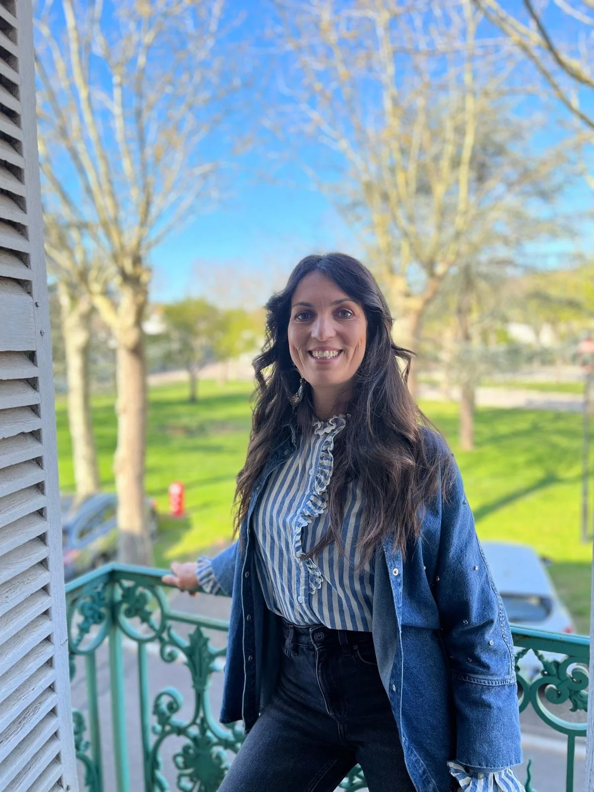 Une femme souriante se tient sur un balcon en bois, avec une balustrade en fer forgé vert, derrière elle un paysage de parc avec des arbres sans feuilles et un ciel bleu clair.