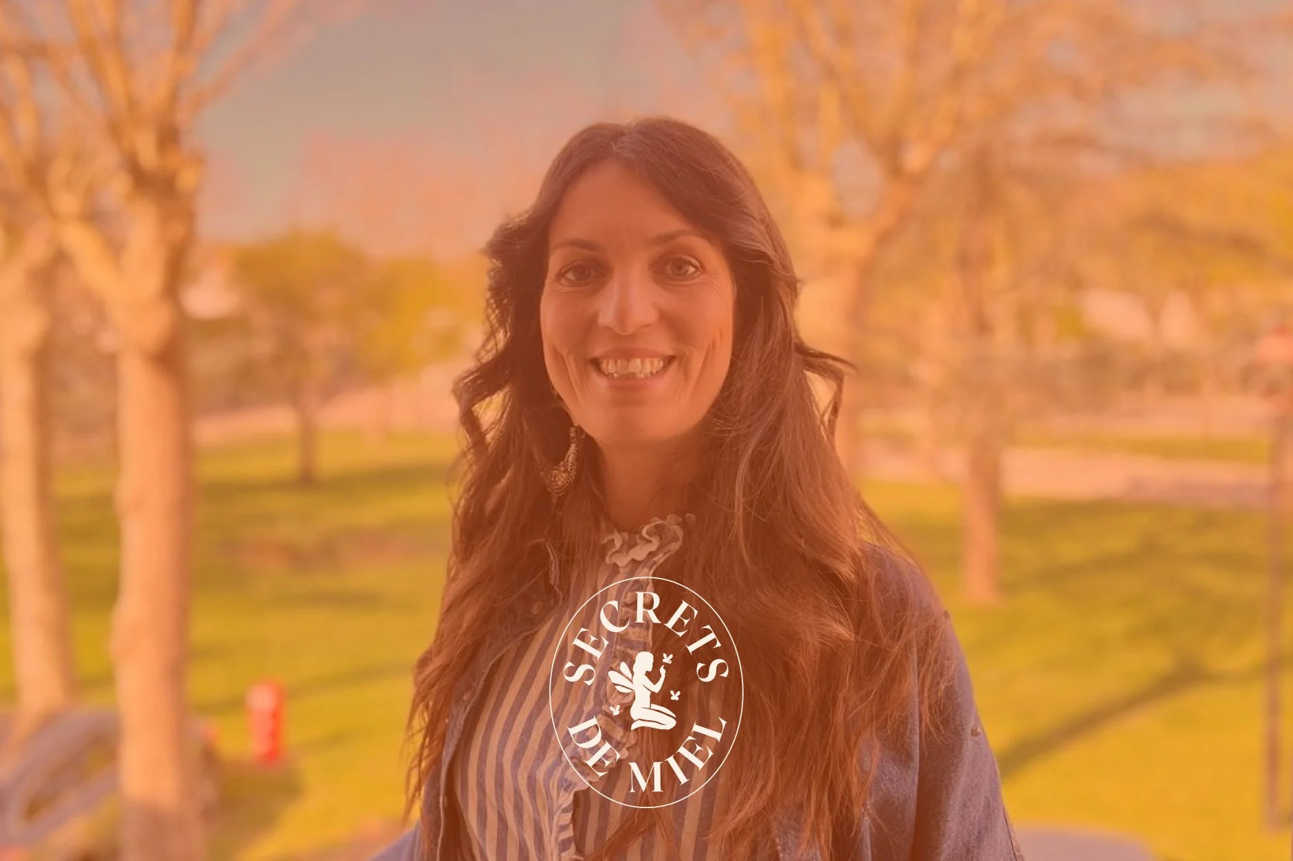 Une femme souriante avec des cheveux longs, portant une veste en cuir et une chemise à rayures, debout dans un parc avec des arbres en automne.