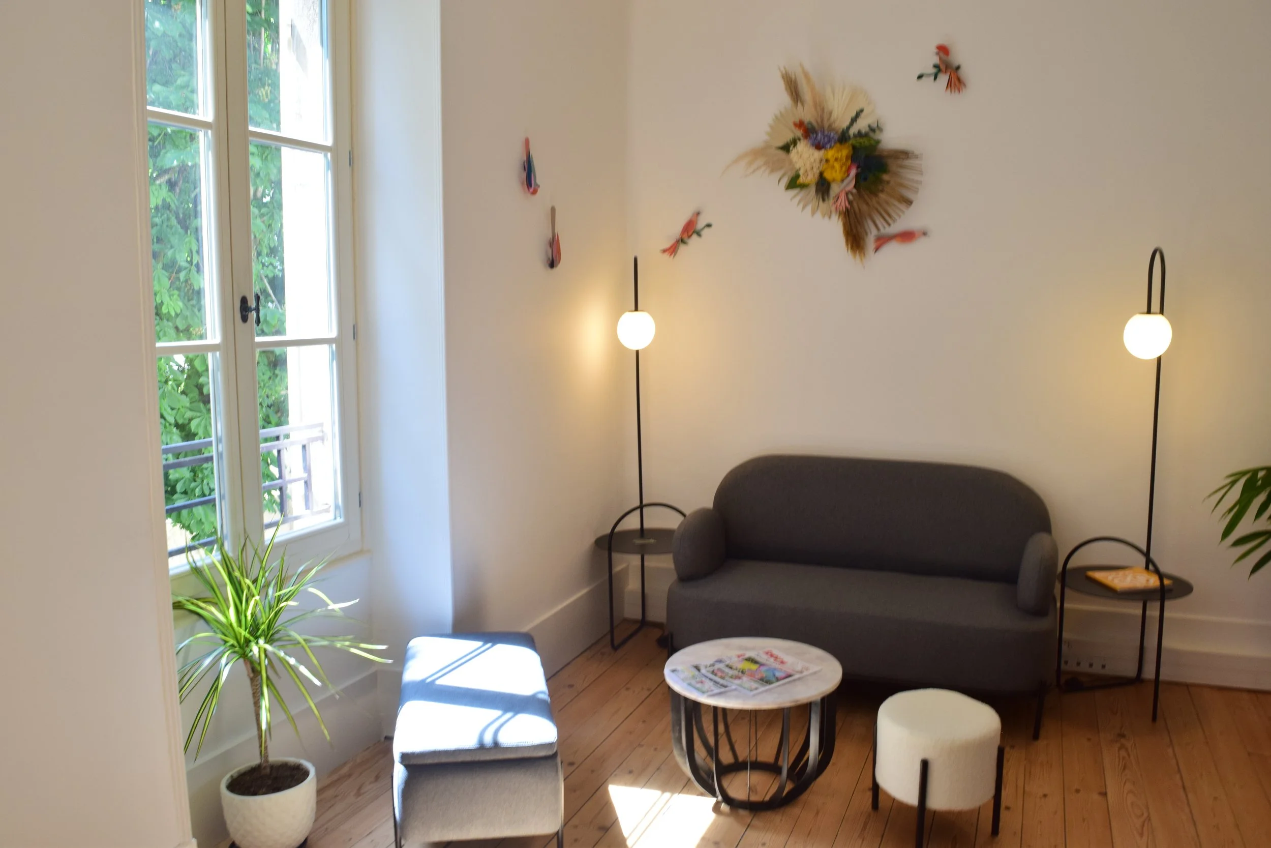 Salon moderne avec un canapé gris, une table ronde en marbre, deux lampes avec abat-jour rond, une plante verte en pot près de la fenêtre, une décoration murale avec des fleurs séchées, et une fenêtre laissant entrer la lumière naturelle.