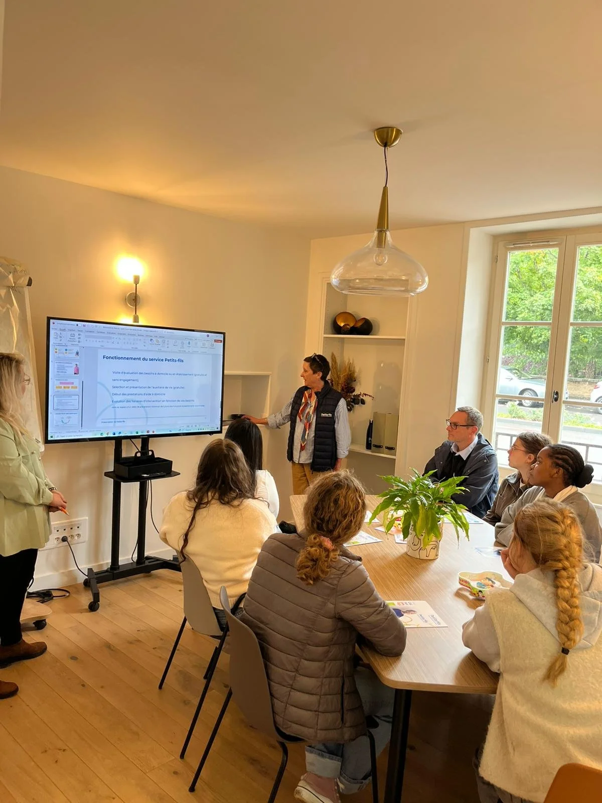Une femme présente une diapositive lors d'une réunion à une table en bois, entourée de plusieurs personnes assises. La pièce est éclairée par la lumière naturelle par une grande fenêtre et une lampe suspendue. On voit une plante verte sur la table et une étagère décorative en arrière-plan.