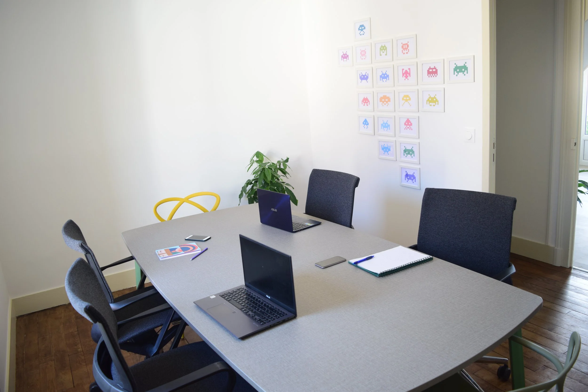 Salle de réunion avec table, plusieurs ordinateurs portables, des chaises, une plante verte et des dessins pixel art de jeux vidéo "Space Invaders" accrochés au mur.