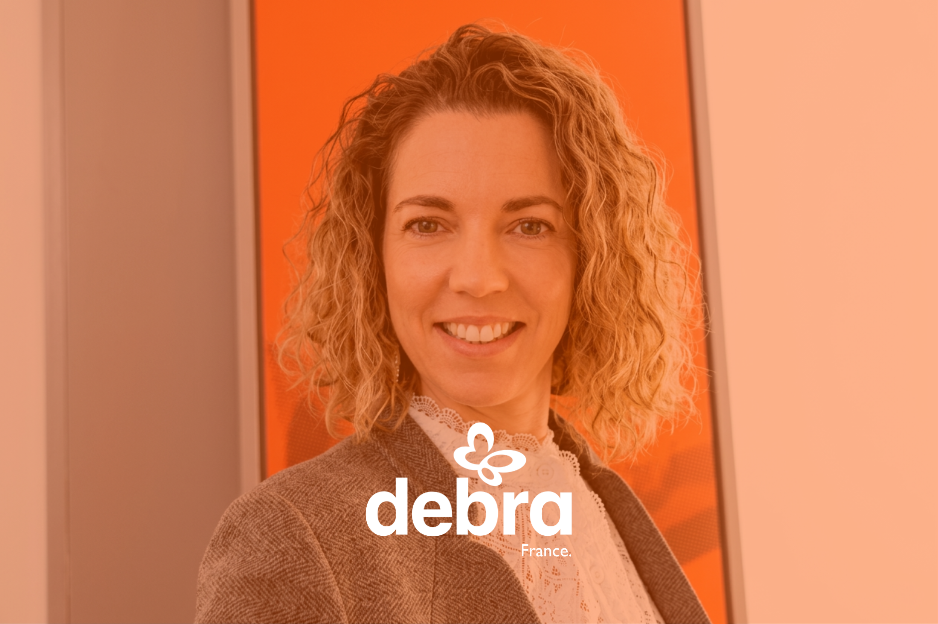 Portrait d'une femme blondie aux cheveux bouclés, souriante, portant un blazer et une chemise, avec un fond orange. Logo de Debra France en bas de l'image.