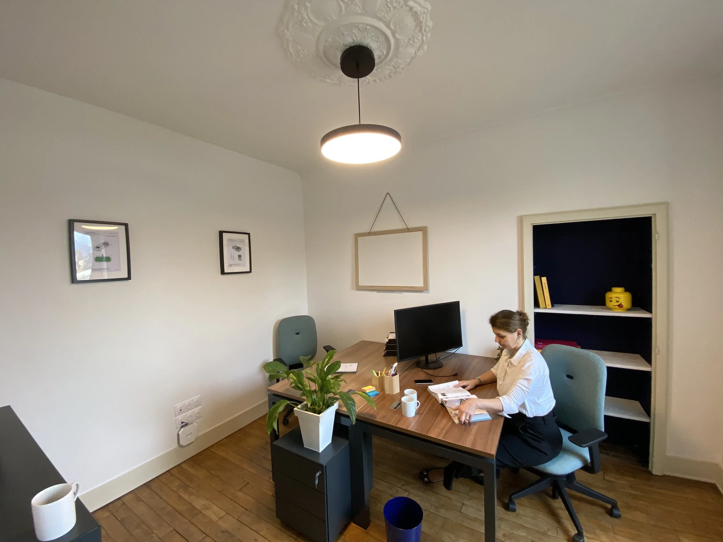 Une femme assise à un bureau en train de lire un magazine ou un livre dans une salle de bureau moderne. Sur le bureau, il y a une plante verte, des tasses, et des fournitures de bureau. La pièce a des murs blancs, un plafond orné de moulures, un écran d'ordinateur, et des cadres accrochés. Un étagère blanche avec des livres et une tête de Lego jaune décorative est visible à l'arrière.