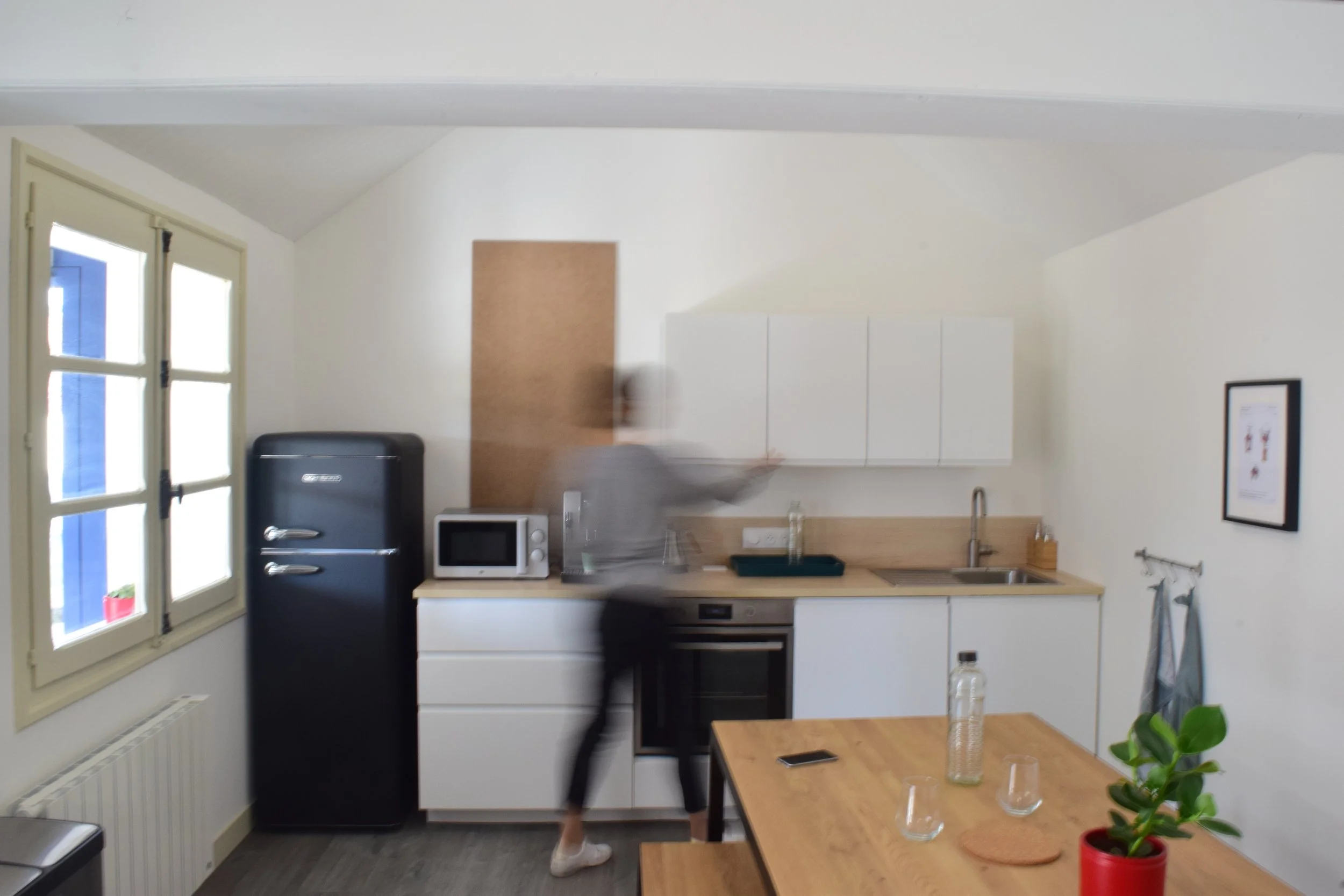 Petite cuisine moderne avec une fenêtre, un réfrigérateur noir, un micro-ondes blanc, un évier, un plan de travail en bois, et une table en bois avec une plante en pot.