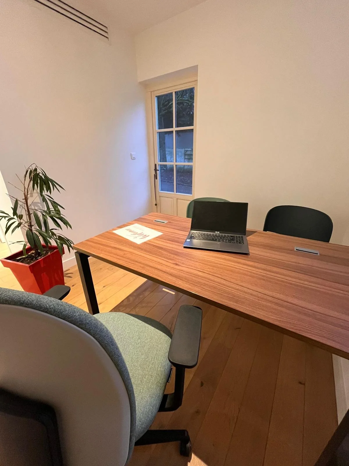 Bureau avec un ordinateur portable, une chaise, une plante en pot rouge, une fenêtre avec une vue extérieure et une porte, murs blancs, sol en bois.