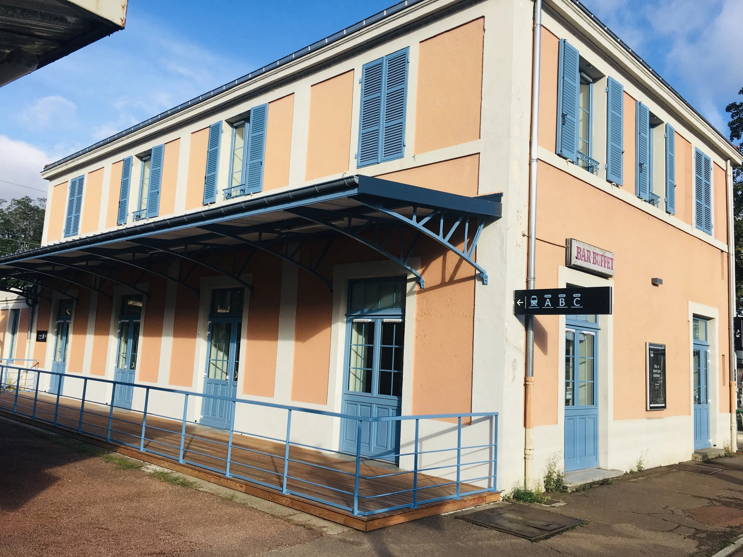 Bâtiment à deux étages en couleur pastel avec volets bleus, signé 'Bar Buffet', avec une rampe en métal bleu entourant une terrasse en bois
