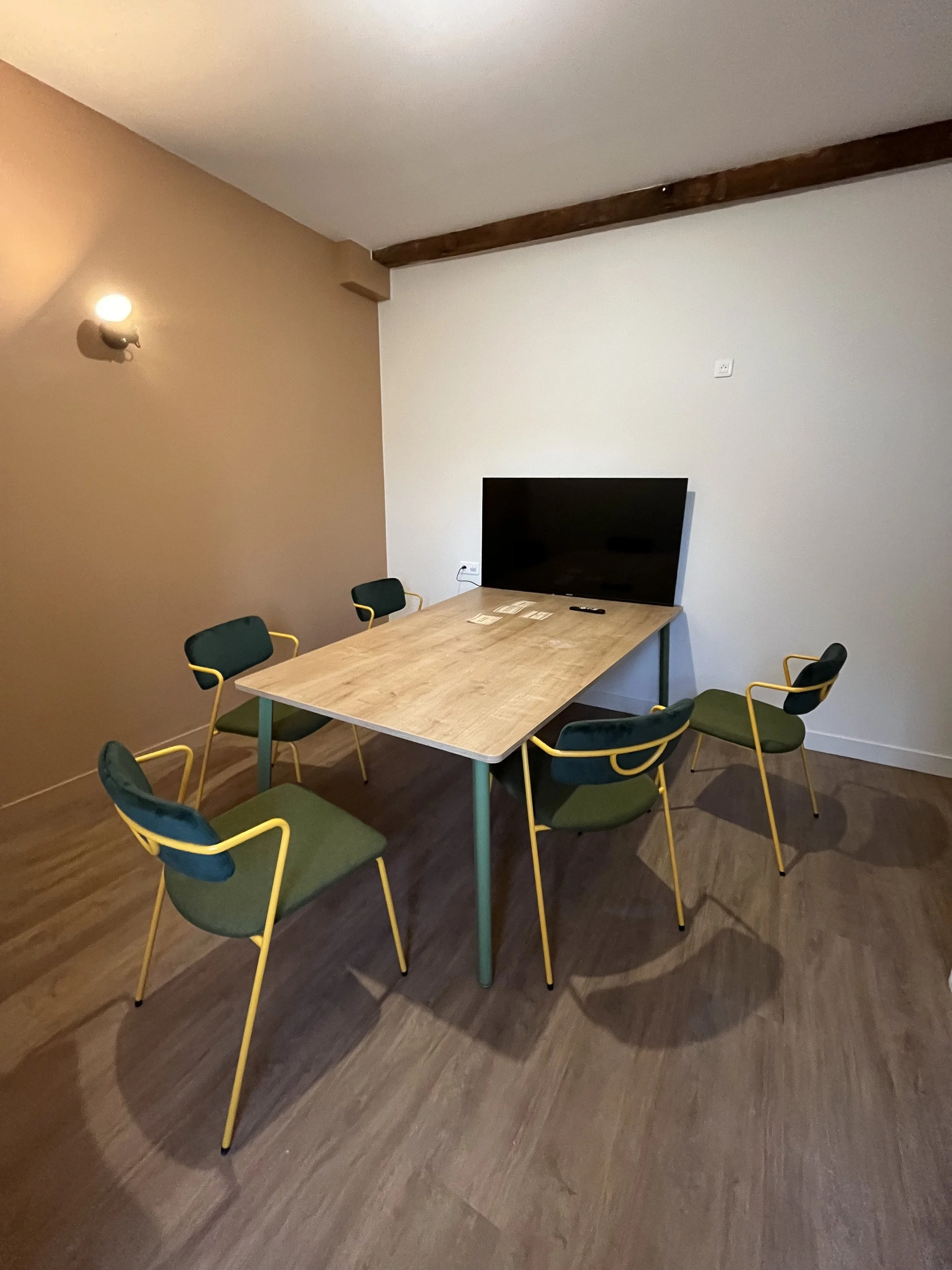 Salle de réunion avec une table en bois, six chaises vertes avec cadres jaunes, un mur blanc, un mur beige, et un mur avec une télévision, lumière murale