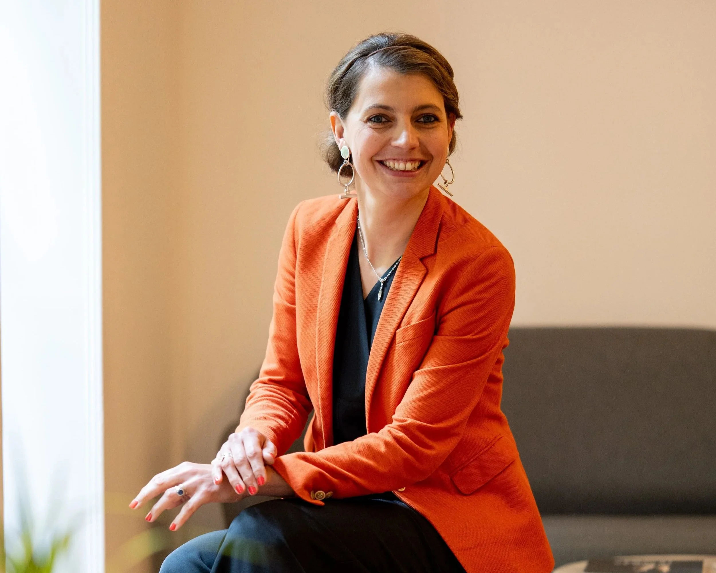 Une femme souriante avec des cheveux courts, portant un blazer orange, des boucles d'oreilles argentées et une broche, est assise à une table dans une pièce avec un mur beige en arrière-plan.