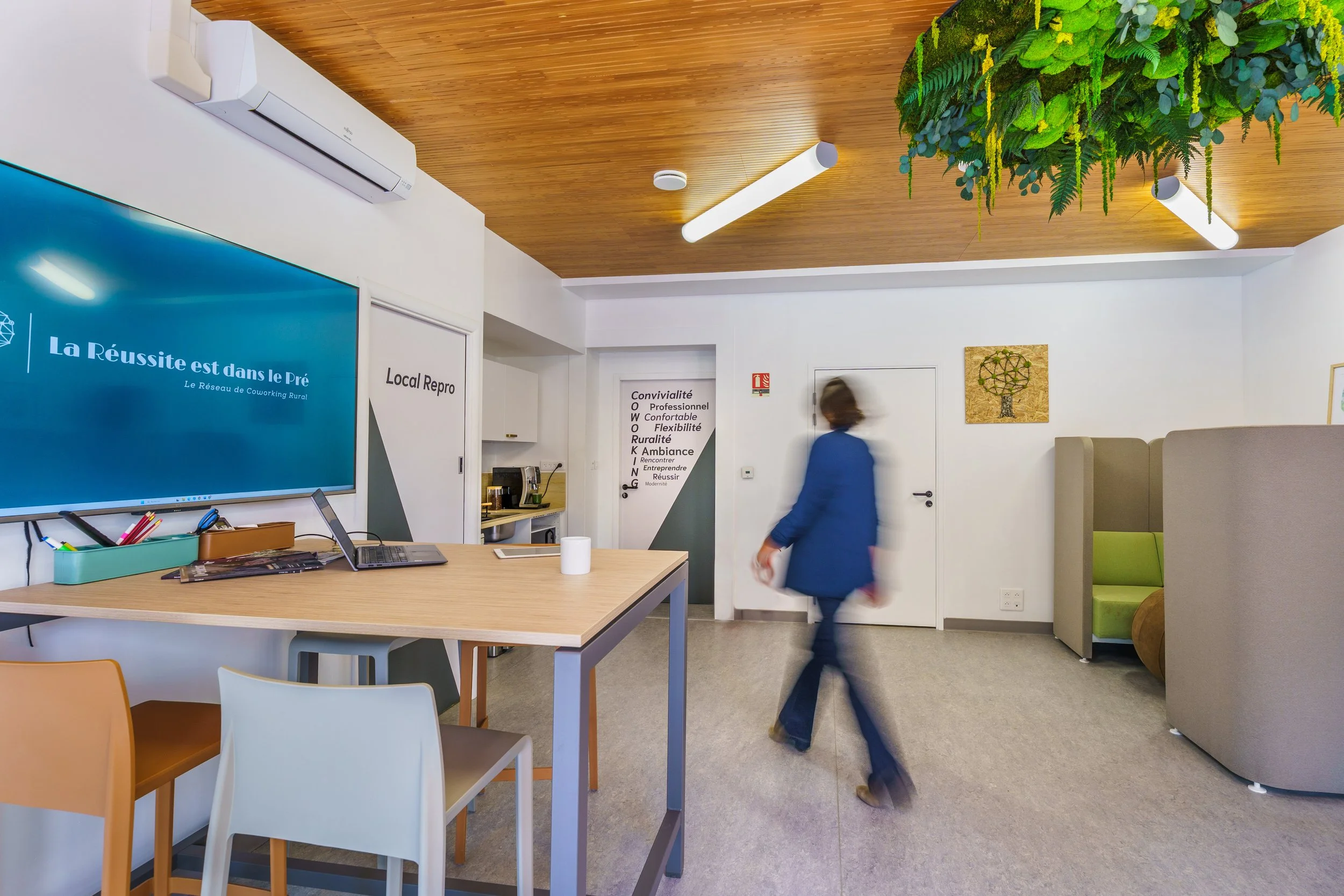 Un espace de coworking moderne avec un plafond en bois, un mur blanc avec des affiches et un grand écran bleu, un comptoir avec un ordinateur portable, une personne en costume marchant à travers la pièce, un arbre vert suspendu au plafond, et un coin salon avec des sièges verts.