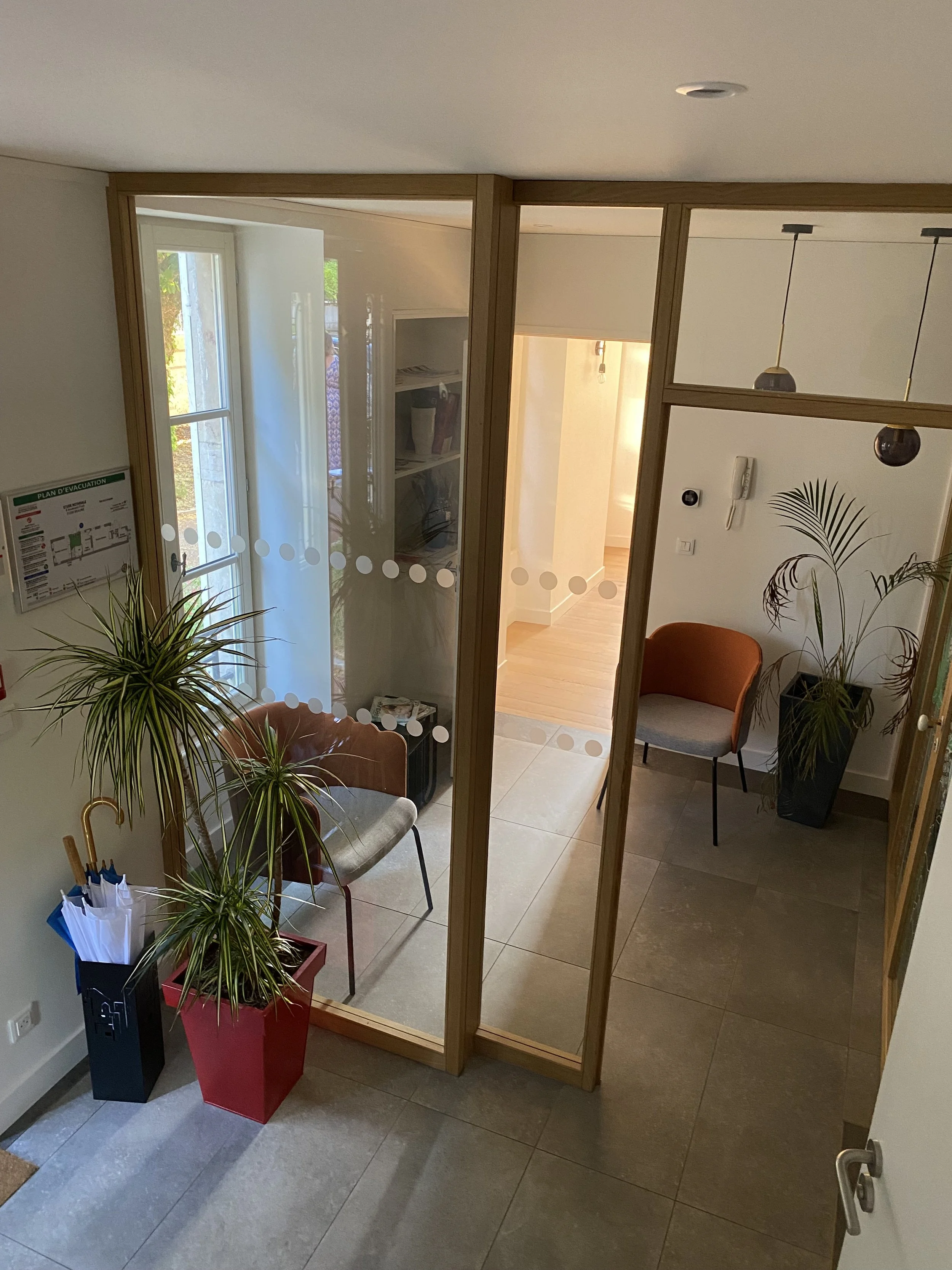 Vue intérieure d'un hall d'entrée séparé par une cloison en bois et verre. Deux plantes en pot, un fauteuil orange, un vestiaire avec un parapluie et un système d'alarme murale visible.