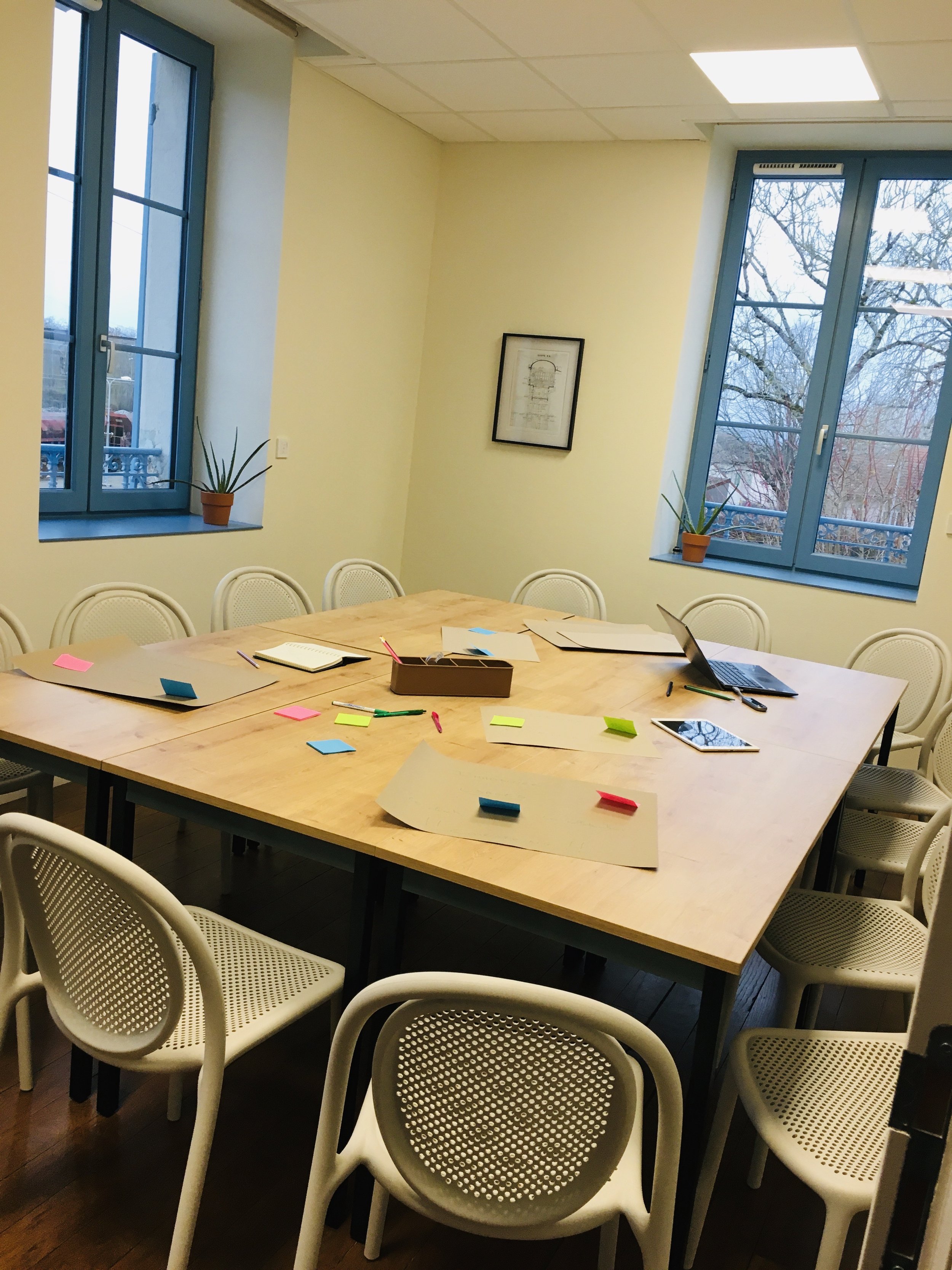 Une salle de réunion avec une grande table en bois, plusieurs chaises en plastique blanc avec des dossiers perforés, des feuilles, des stylos, un ordinateur portable et des notes autocollantes colorées sur la table. Deux fenêtres avec des volets bleus laissent entrer la lumière et offrent une vue sur des arbres nus. La pièce a un plafond similaire à un plafond suspendu et une peinture claire.