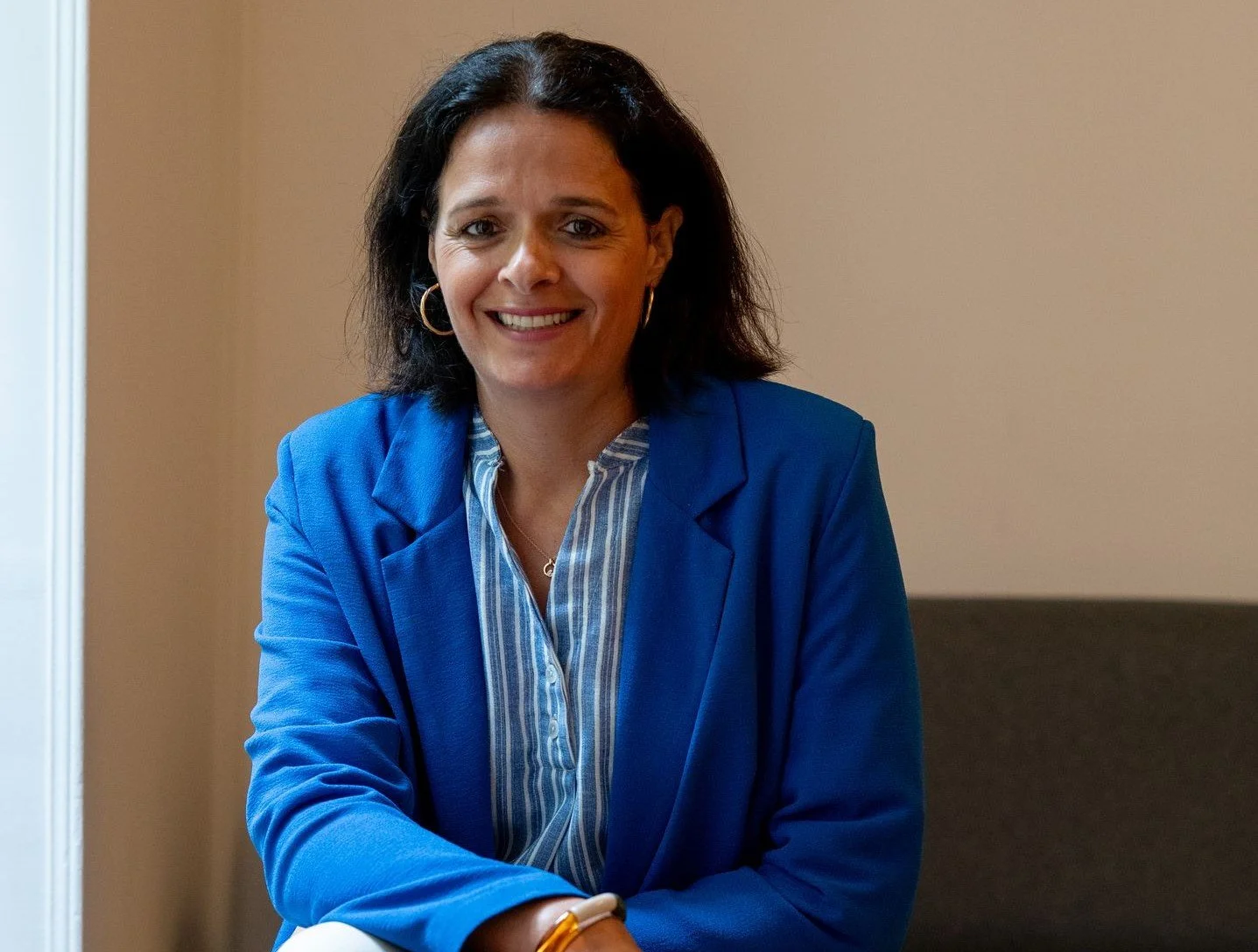 Femme souriante avec des cheveux courts, portant un blazer bleu et une chemise à rayures, assise dans un bureau.