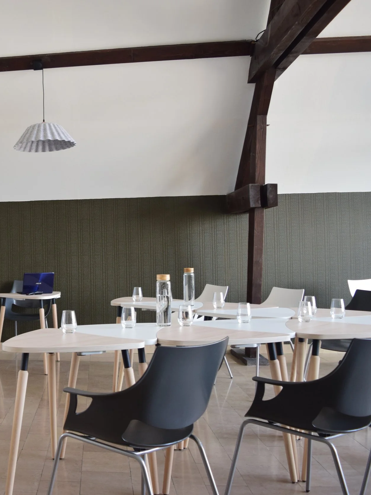 Salle à manger ou salle de réunion avec plusieurs tables rondes, des chaises en plastique, quelques bouteilles d'eau et verres, une lumière suspendue et une décoration épurée avec un mur en tapis vert et des poutres en bois apparentes au plafond.