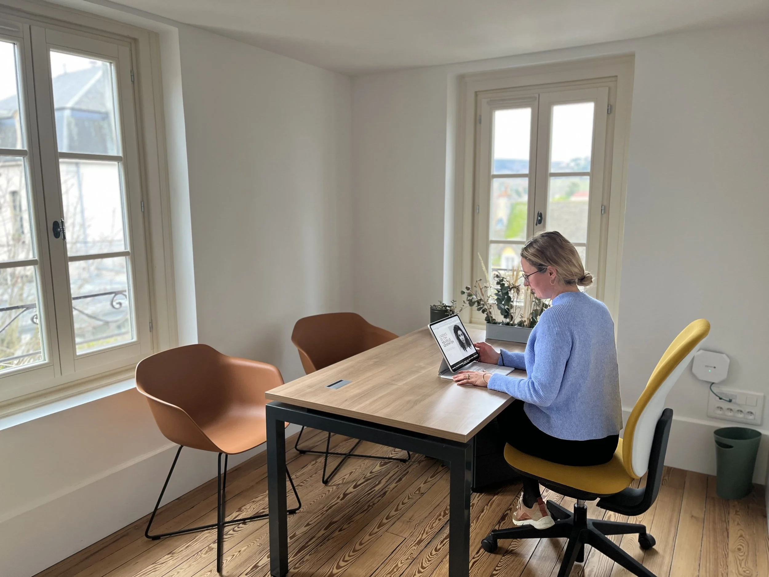 Une femme assise à un bureau, utilisant un ordinateur portable, dans une pièce lumineuse avec deux grandes fenêtres et des chaises de différentes couleurs.