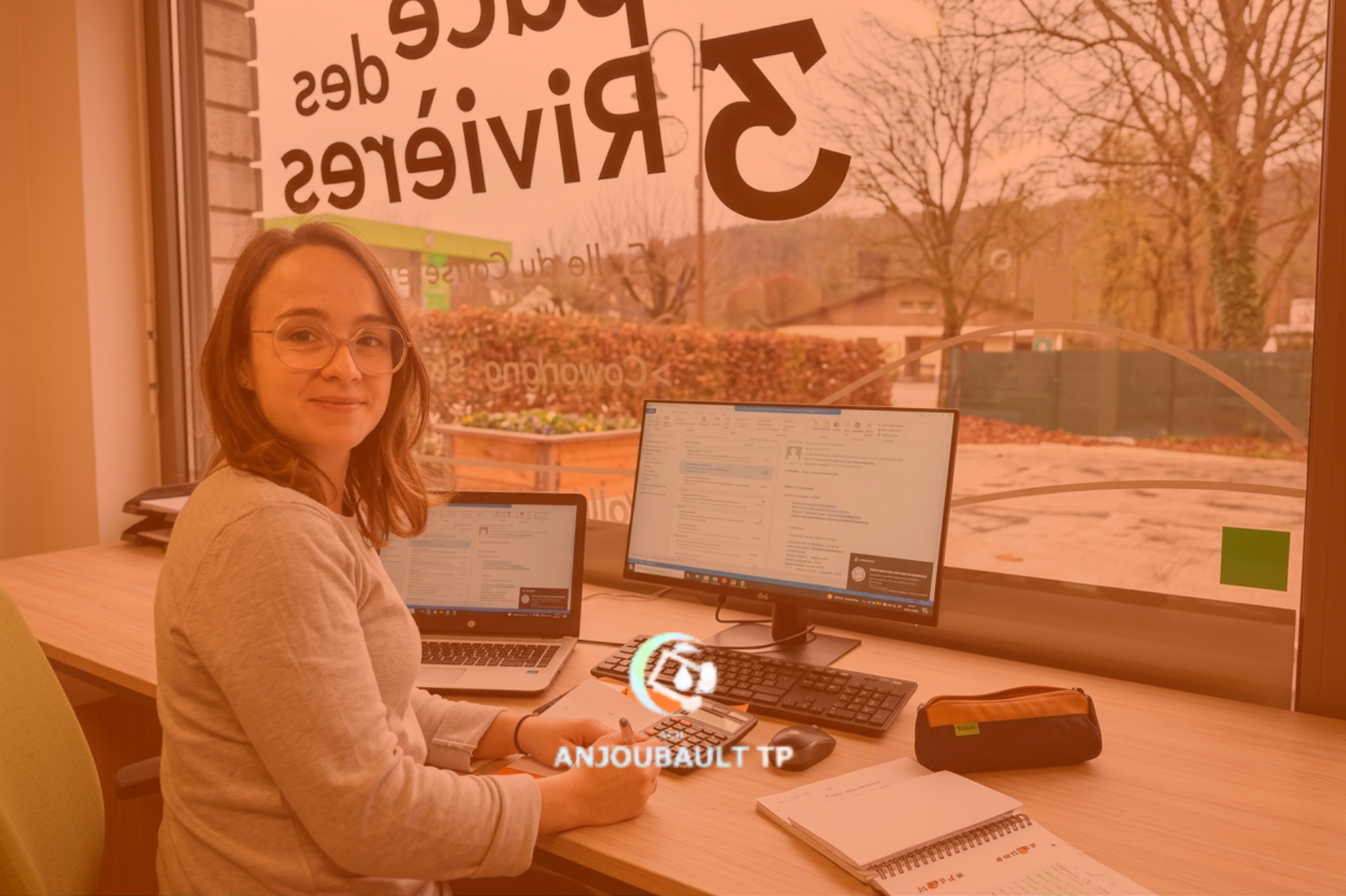 Une femme assise à un bureau avec deux ordinateurs portables, tenant un stylo et un carnet, dans une pièce avec une grande fenêtre. La fenêtre montre un paysage d'automne avec des arbres dénudés et un ciel gris. Sur la vitre, des inscriptions en français et un logo sont visibles.