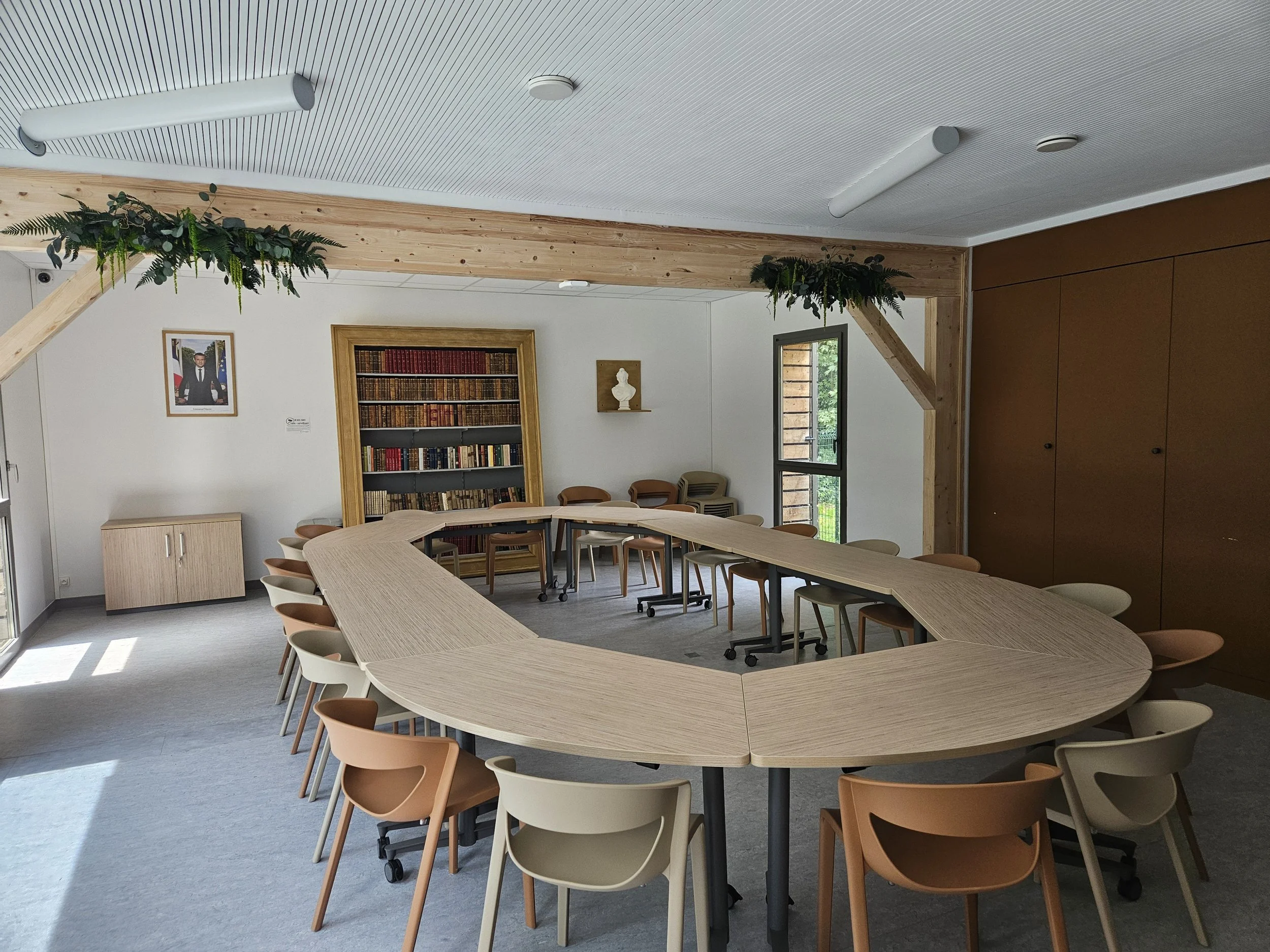 Salle de réunion avec une table en forme de U, chaises en bois, étagère à livres, portrait encadré, petite sculpture, fenêtre donnant sur l'extérieur, décor simple avec un mélange de bois clair et blanc.