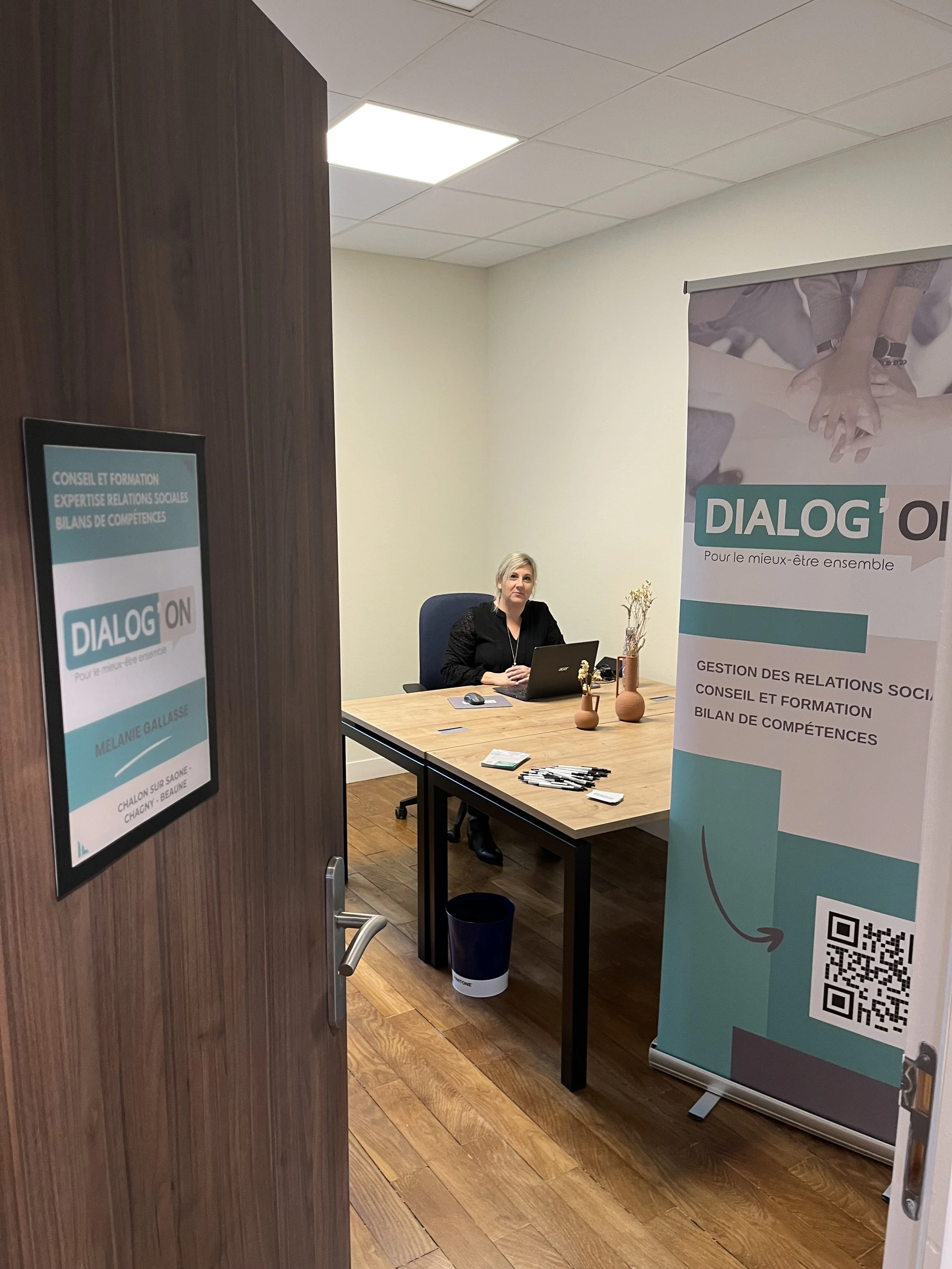 Une femme assise à un bureau dans une salle de réunion ou un bureau, avec un panneau d'affichage et une banderole promotionnelle à proximité.
