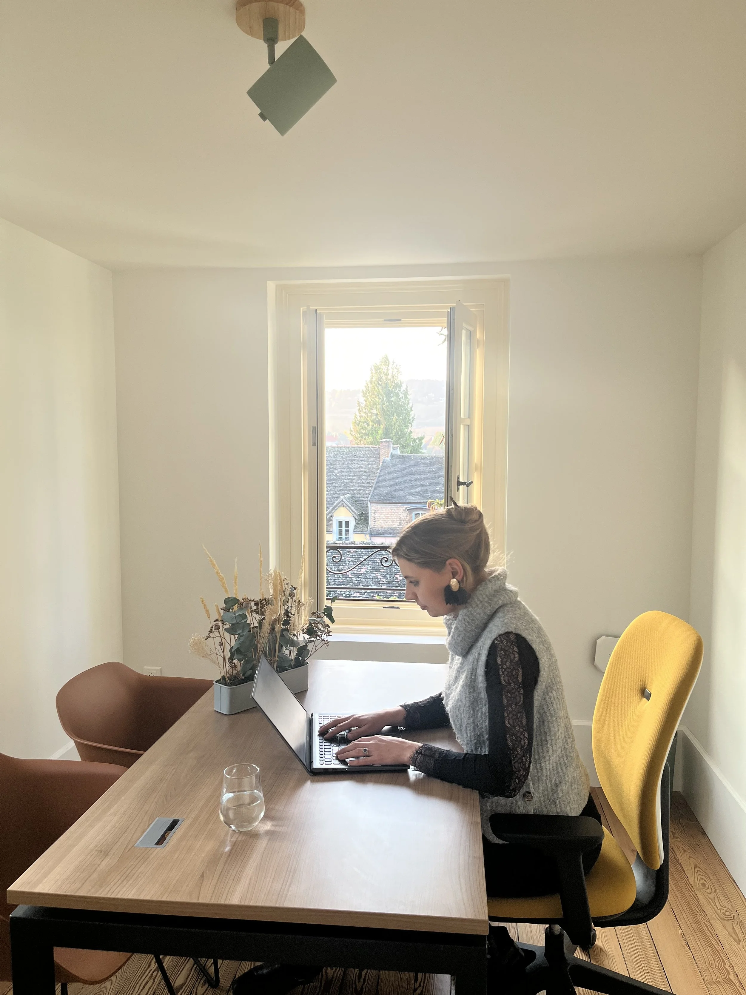 Femme assise à une table de bureau, utilisant un ordinateur portable, dans une pièce lumineuse avec fenêtre ouverte laissant entrer la lumière naturelle, décorée avec un vase de fleurs et un fauteuil marron.