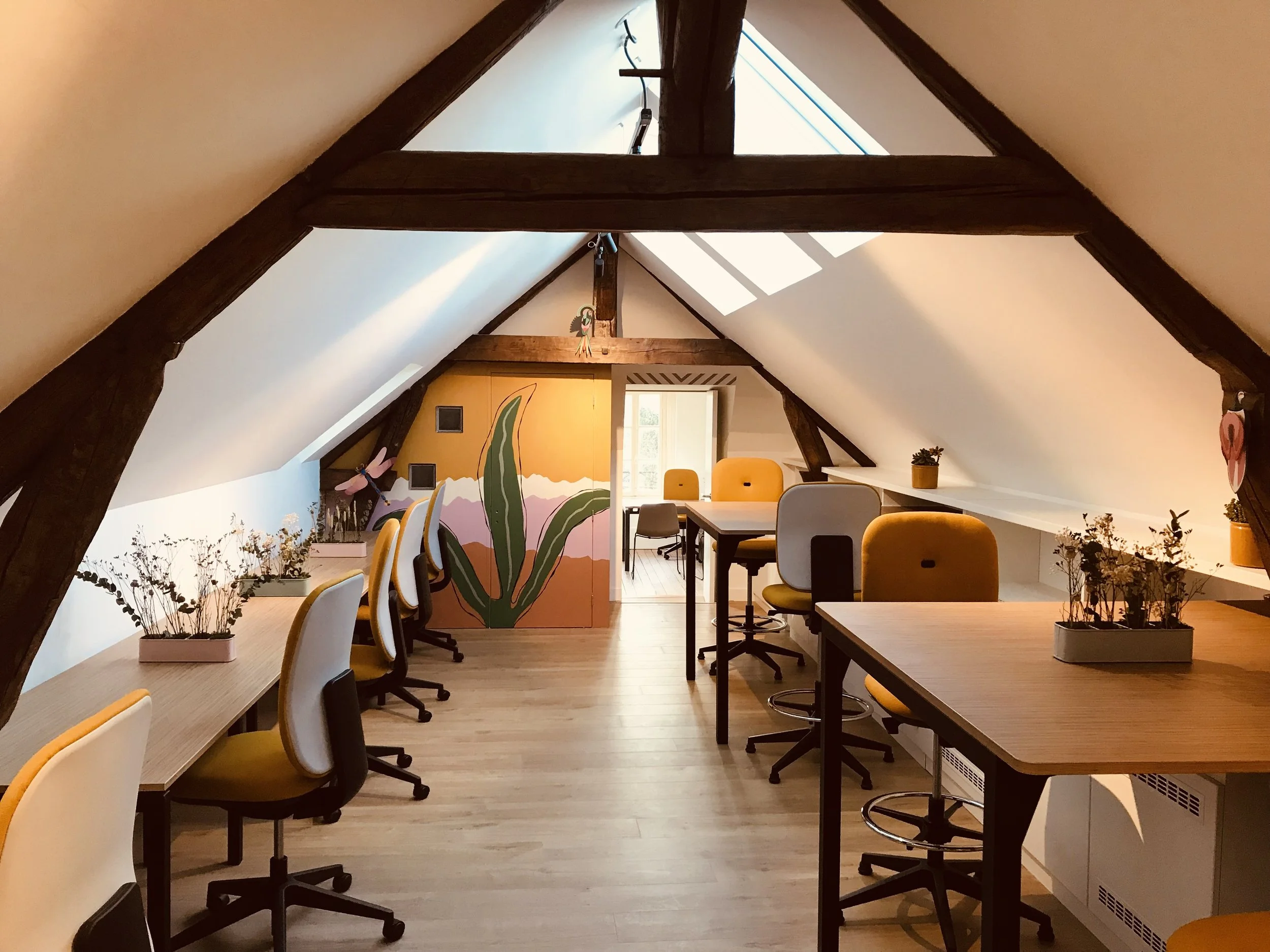 Une salle de réunion dans un loft avec poutres en bois apparentes, mobilier lumineux et décoration végétale. Un mur peint avec une scène de champs de cactus et montagnes en arrière-plan.