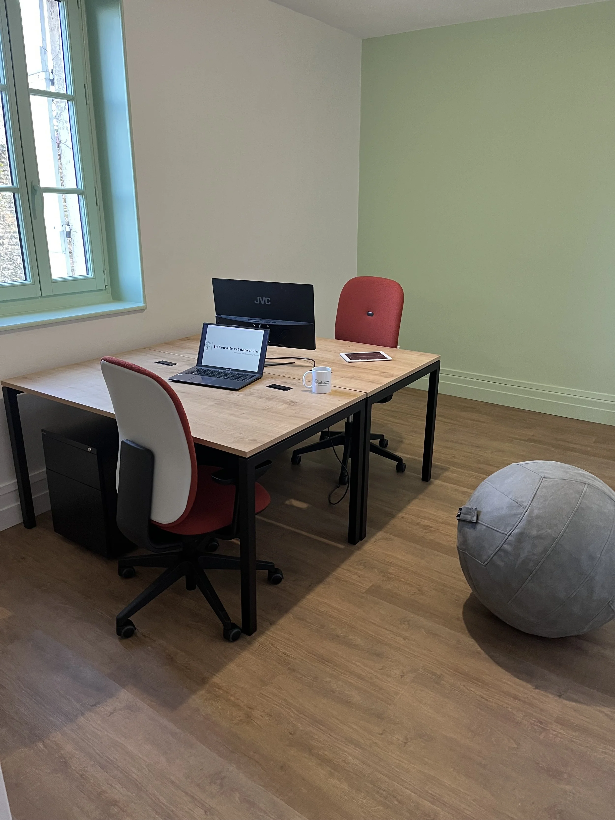 Bureau avec deux chaises, un ordinateur portable, un moniteur, une tasse, une tablette et un ballon de fitness dans une pièce aux murs verts et blancs, avec un sol en bois et une fenêtre donnant sur l'extérieur.