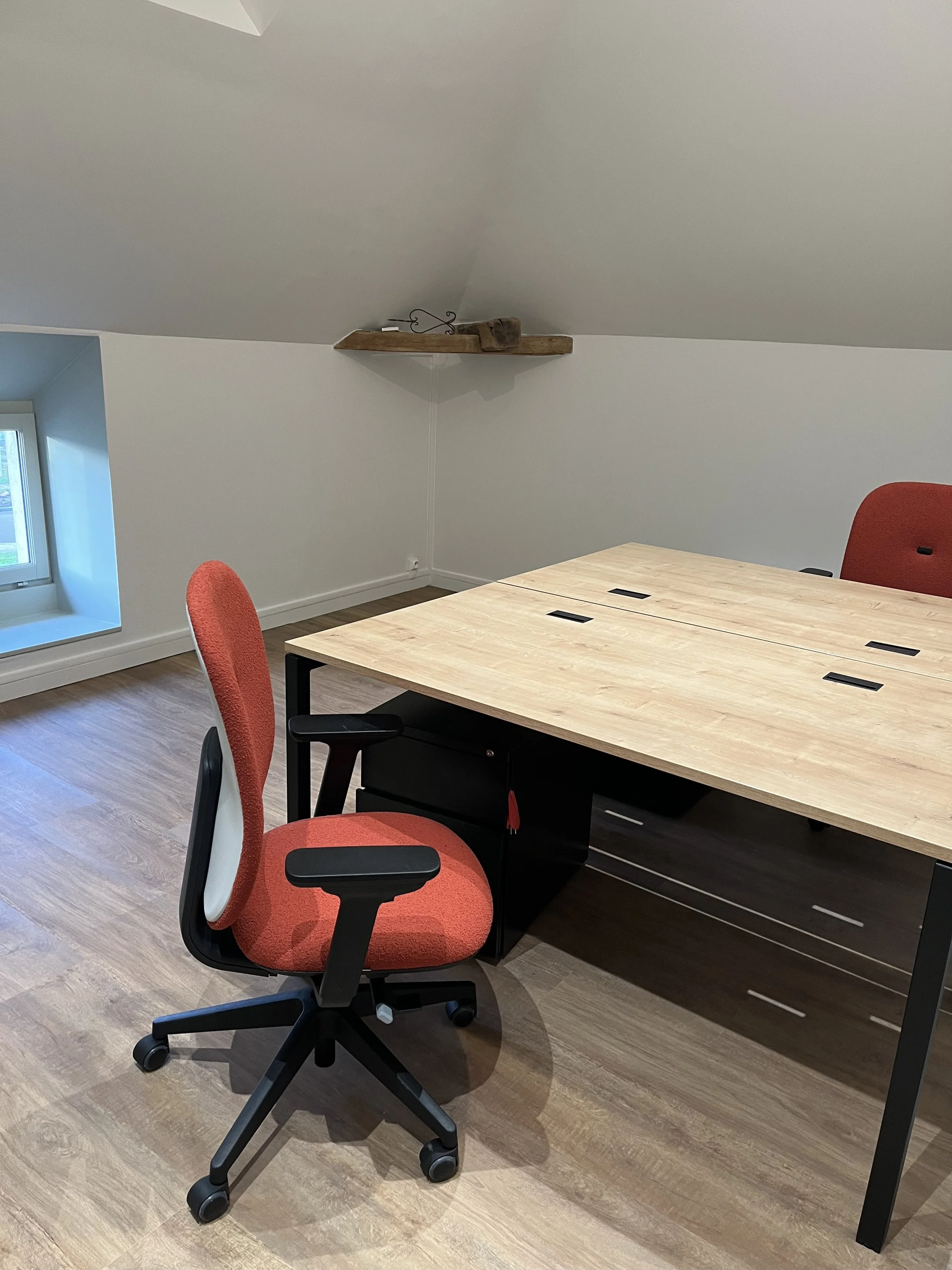 Bureau avec une table en bois et une chaise de bureau rouge, dans une pièce avec sol en bois clair et mur blanc, sous une poutre en bois.