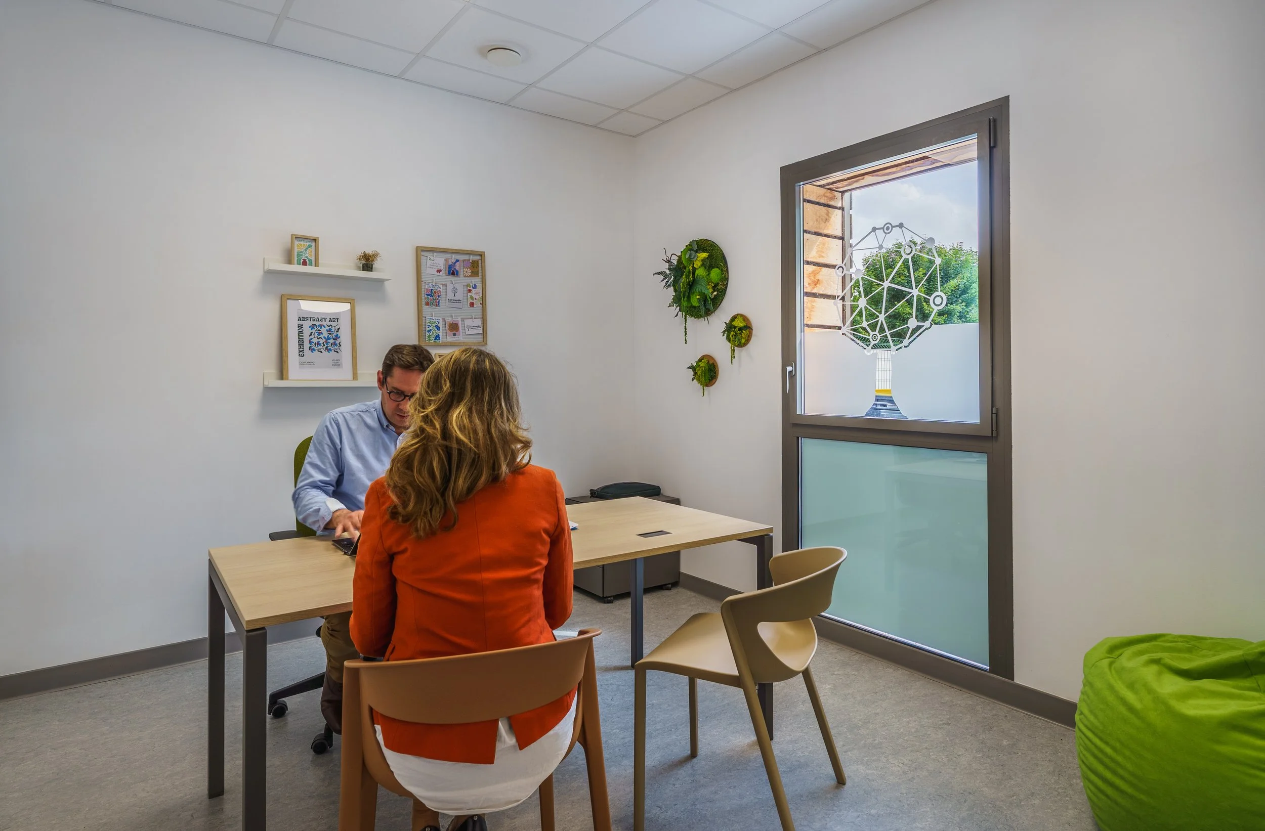 Trois personnes en réunion dans une salle de bureau, deux assises face à face, une autre face de dos. La pièce a une fenêtre avec un décor suspendu en forme de sphère géométrique et des plantes murales vertes.