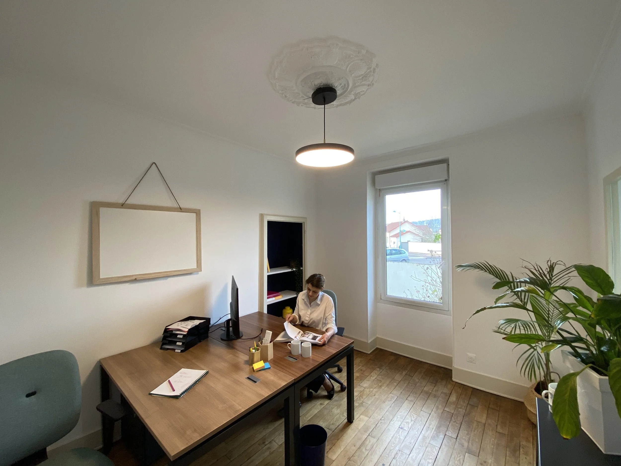 Une personne assise à un bureau en train de lire un magazine dans un bureau lumineux avec une grande fenêtre, des plantes et des décorations murales.