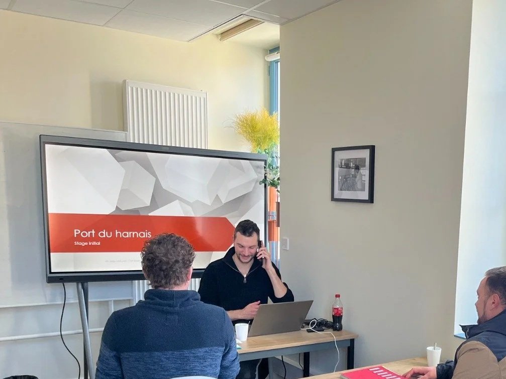 Une salle de réunion avec trois personnes. Une personne au centre utilise un ordinateur portable et parle au téléphone. Le grand écran derrière affiche 'Port du harnais' et 'Stage initial'.
