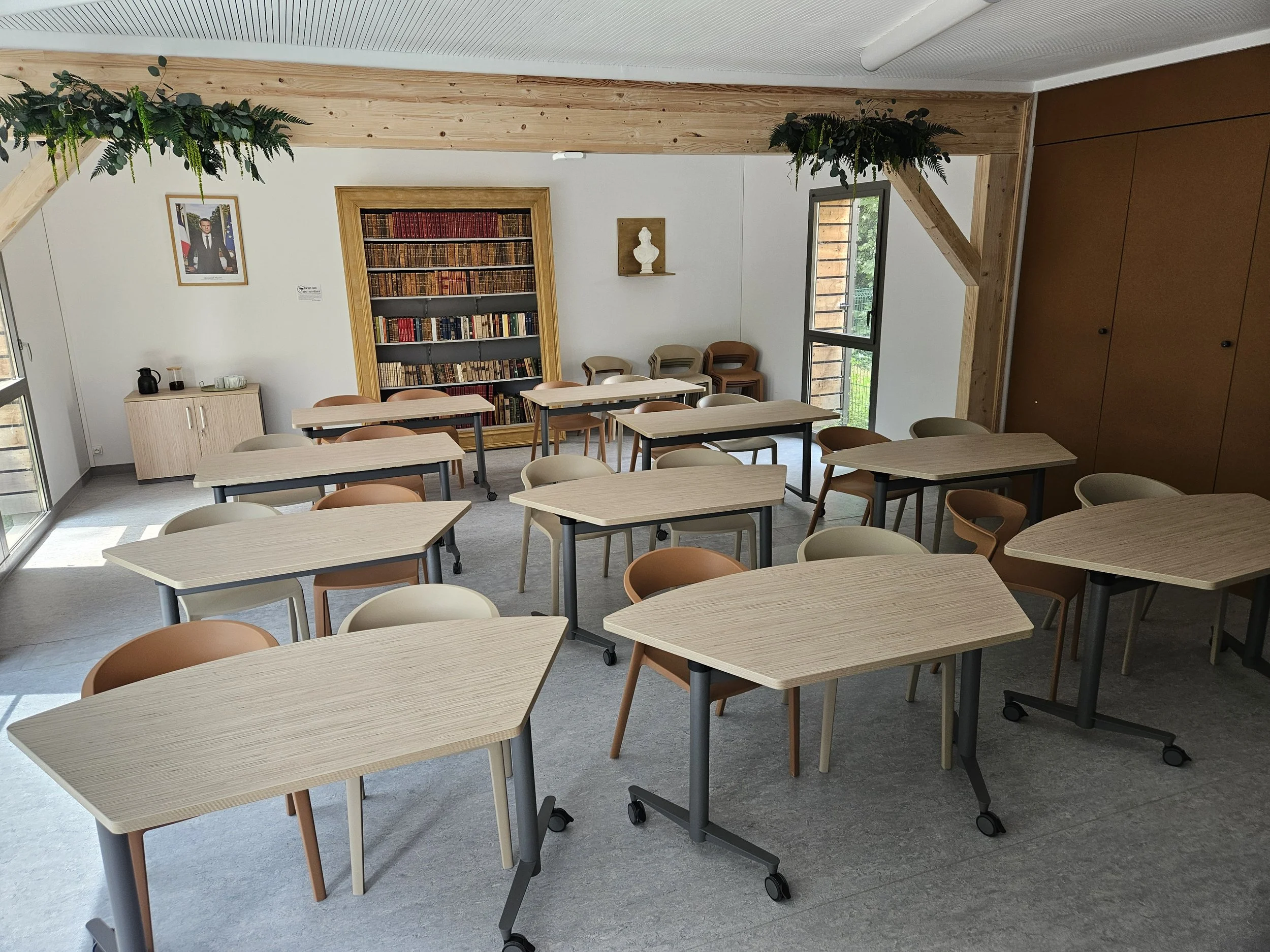Une salle de classe lumineuse avec plusieurs tables en bois clair et des chaises colorées. Il y a une bibliothèque murale remplie de livres, une fenêtre laissant entrer la lumière naturelle, et des décorations végétales suspendues au plafond.