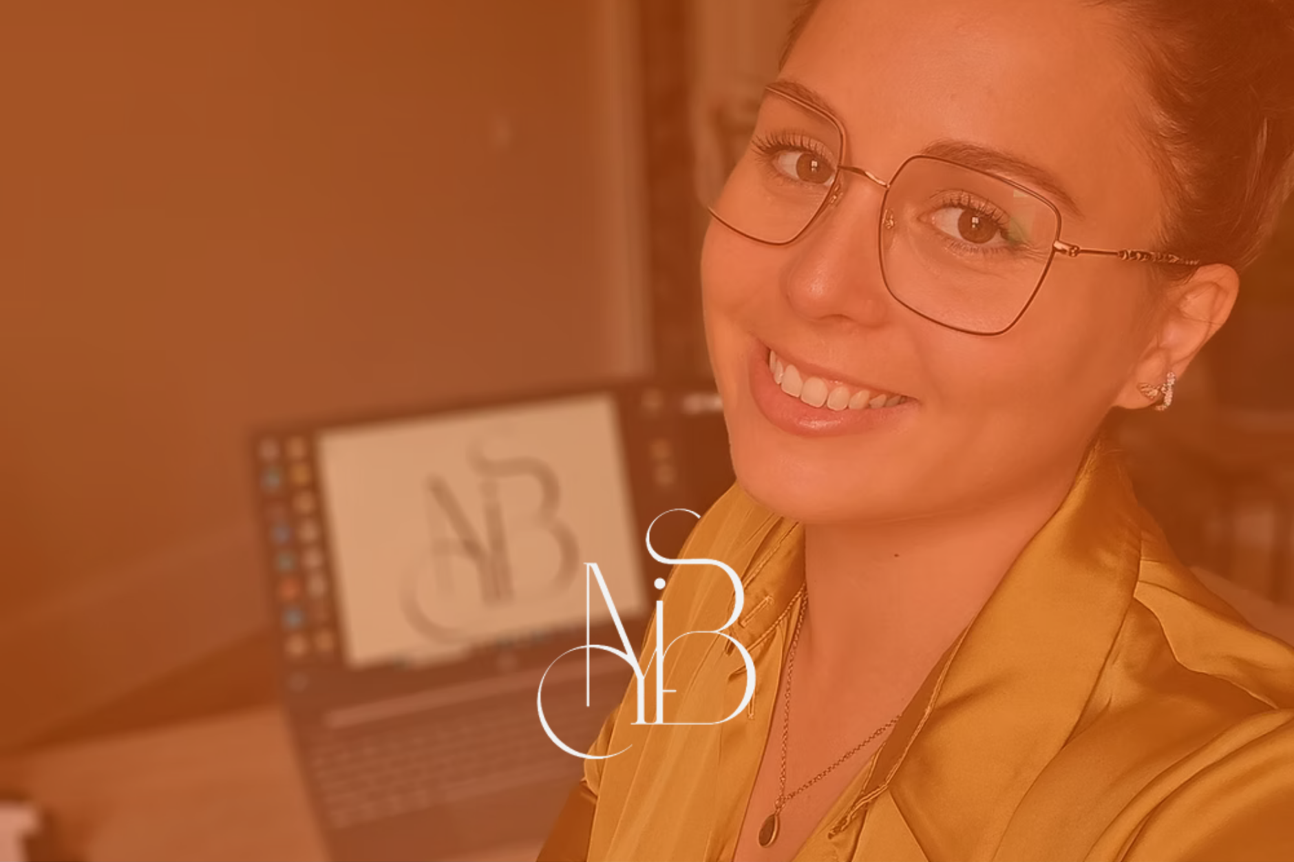 Une femme souriante portant des lunettes et un haut en satin jaune, en arrière-plan un ordinateur. La photo a une teinte orangée.