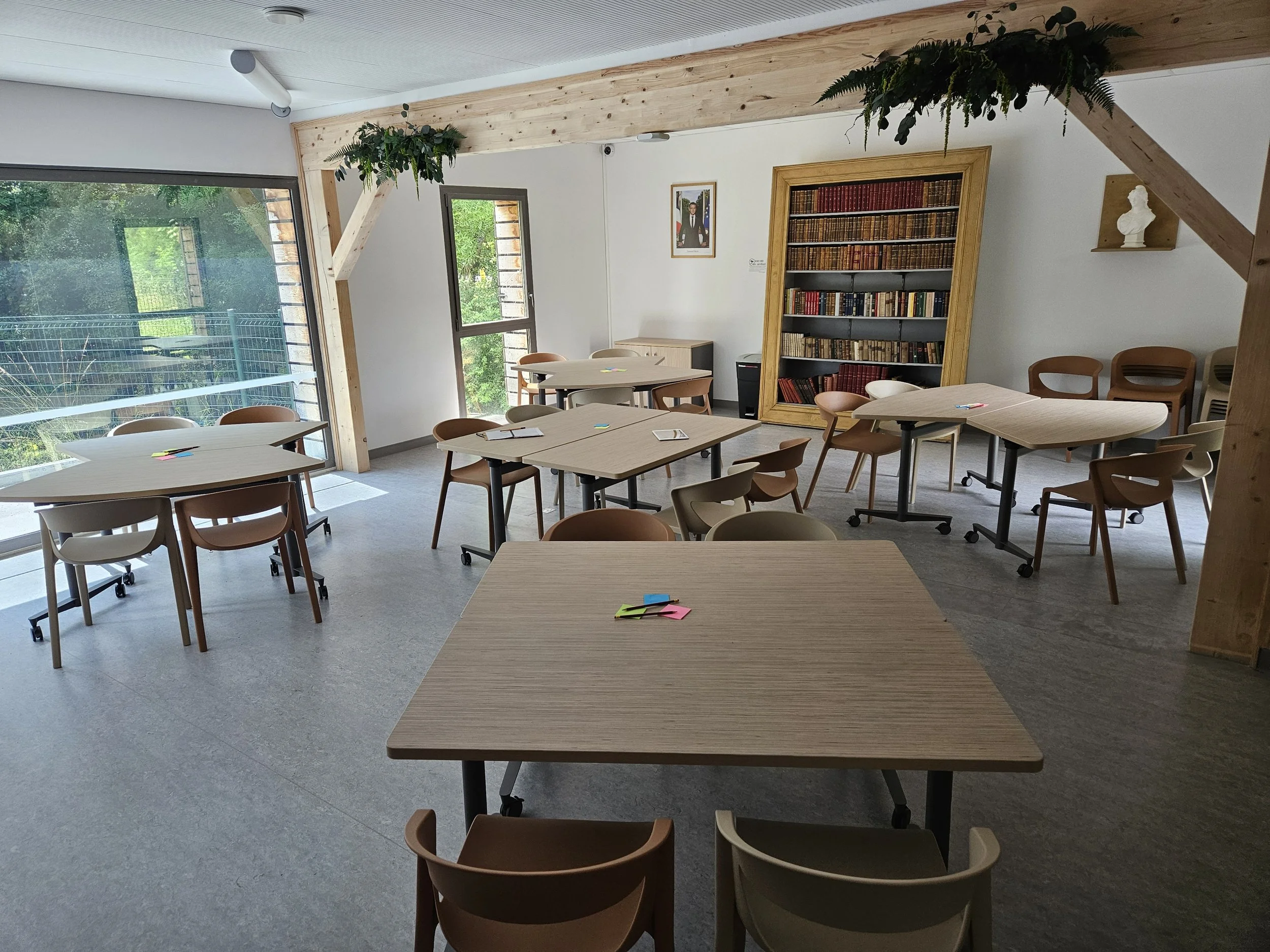 Salle de réunion avec tables rondes, chaises en bois, bibliothèque ancienne avec livres, fenêtres laissant entrer de la lumière naturelle, décor épuré avec plantes vertes suspendues.