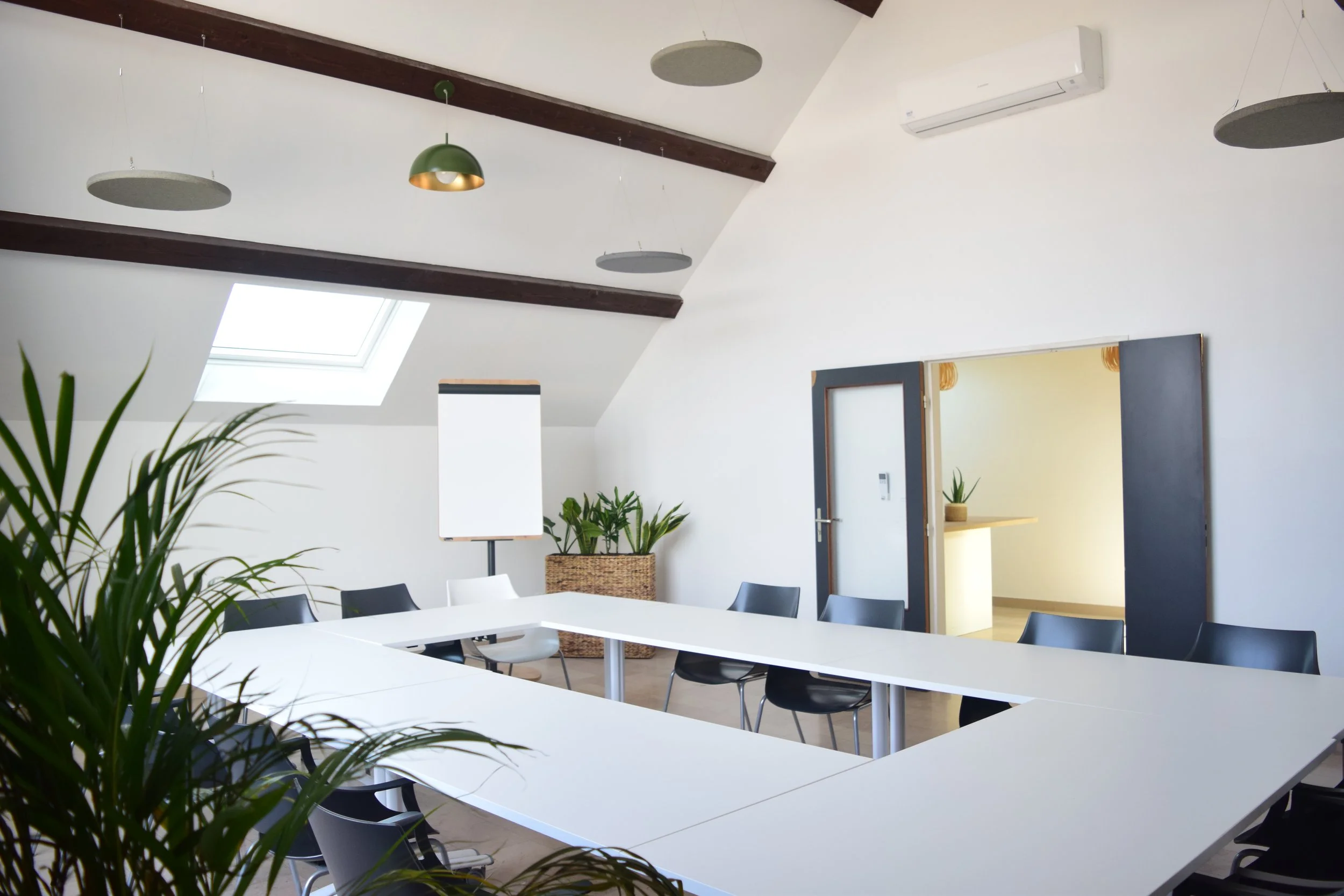 Une salle de réunion moderne avec de grandes tables blanches, des chaises noires, des plantes vertes, des luminaires suspendus et une fenêtre de plafond laissant entrer la lumière naturelle.