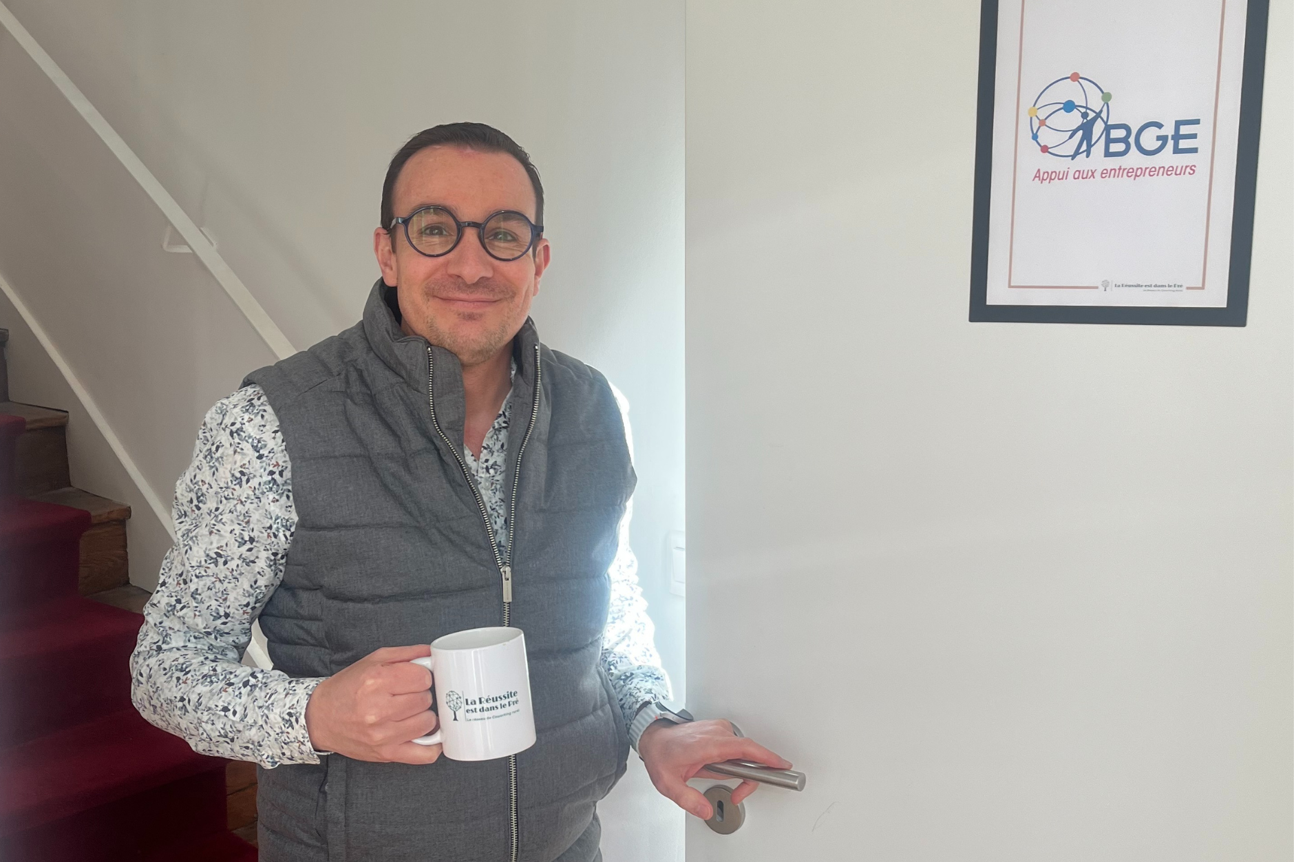 Homme souriant portant une veste en gilet gris et un chemisier à imprimé floral, tenant une tasse blanche avec l'inscription "la Réussite est dans le pré", à côté d'un panneau avec le logo de BGE.