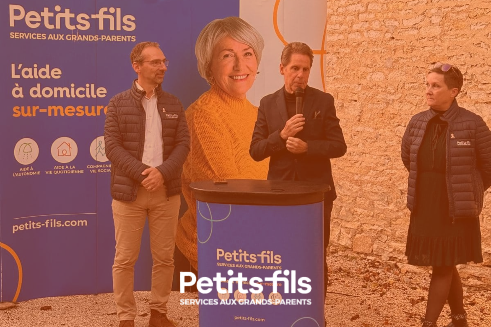 Une affiche promotionnelle pour Petits-fils, un service d’aide à domicile pour les grands-parents, avec trois personnes parlant lors d’un événement, devant un mur en pierre, avec un logo et un slogan sur la bannière.
