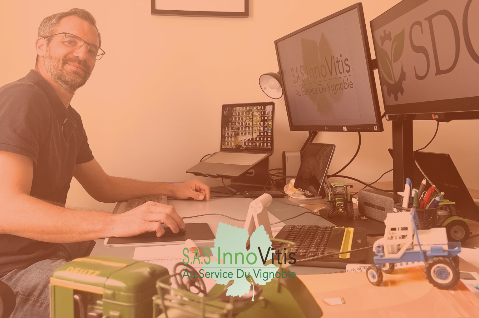Un homme assis à un bureau avec deux écrans d'ordinateur et des objets de bureau, au service du vignoble, avec un logo «SAS InnoVitis» affiché sur un écran et une inscription «Au Service Du Vignoble»