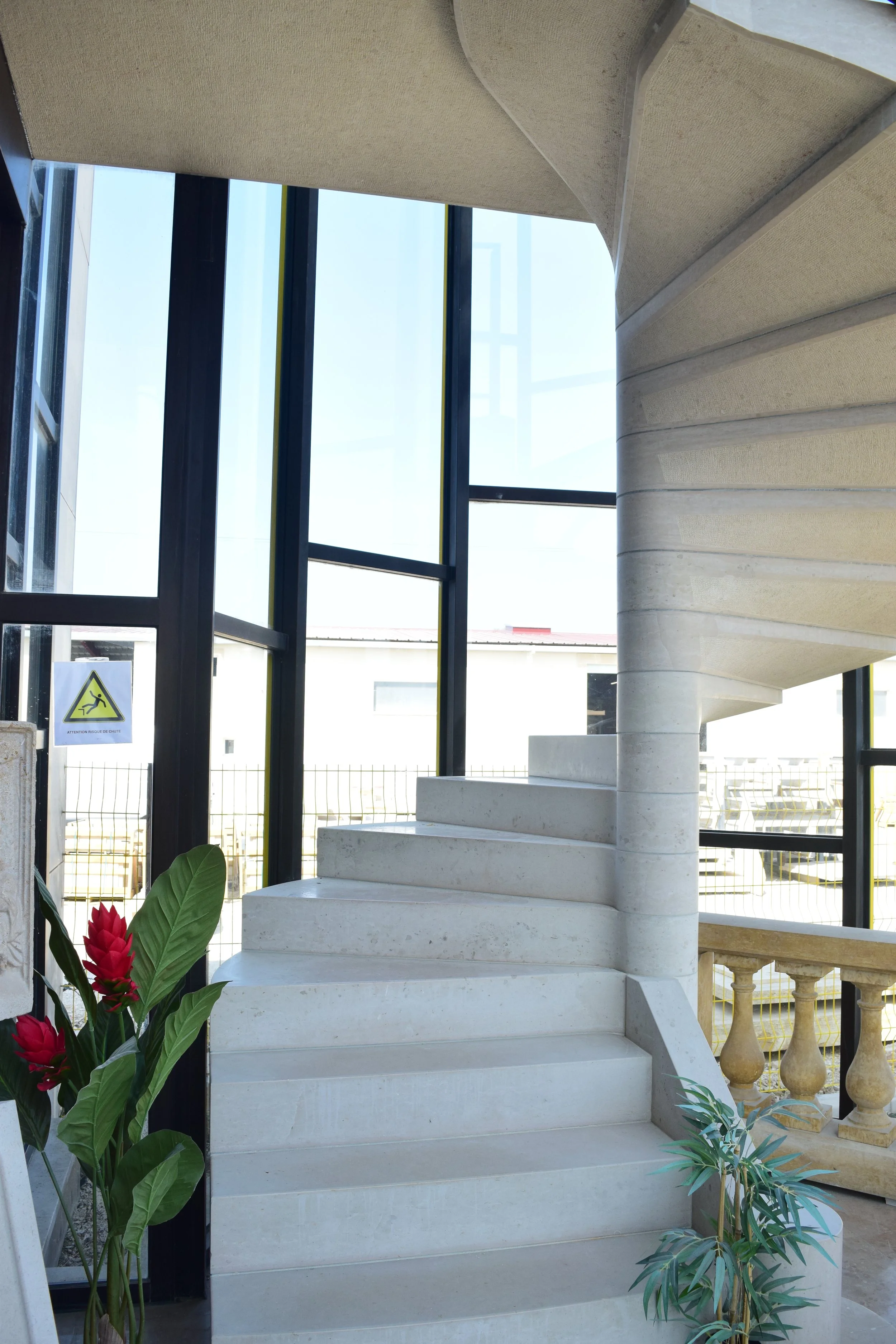 Escalier en pierre blanc avec vague design, entouré de plantes vertes, dans un bâtiment moderne avec des murs en verre.