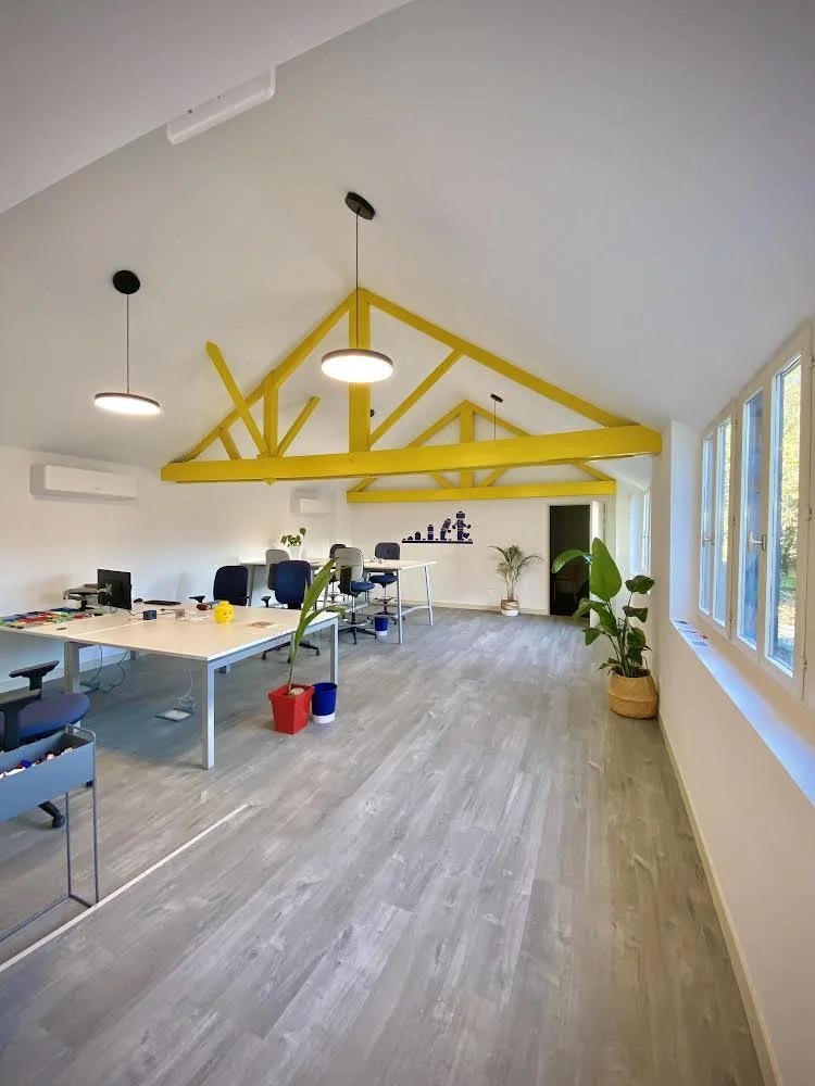 Bureau lumineux avec une grande table de travail, plusieurs chaises, plantes en pot, et une structure en bois peinte en jaune au plafond, avec des fenêtres laissant entrer la lumière naturelle.