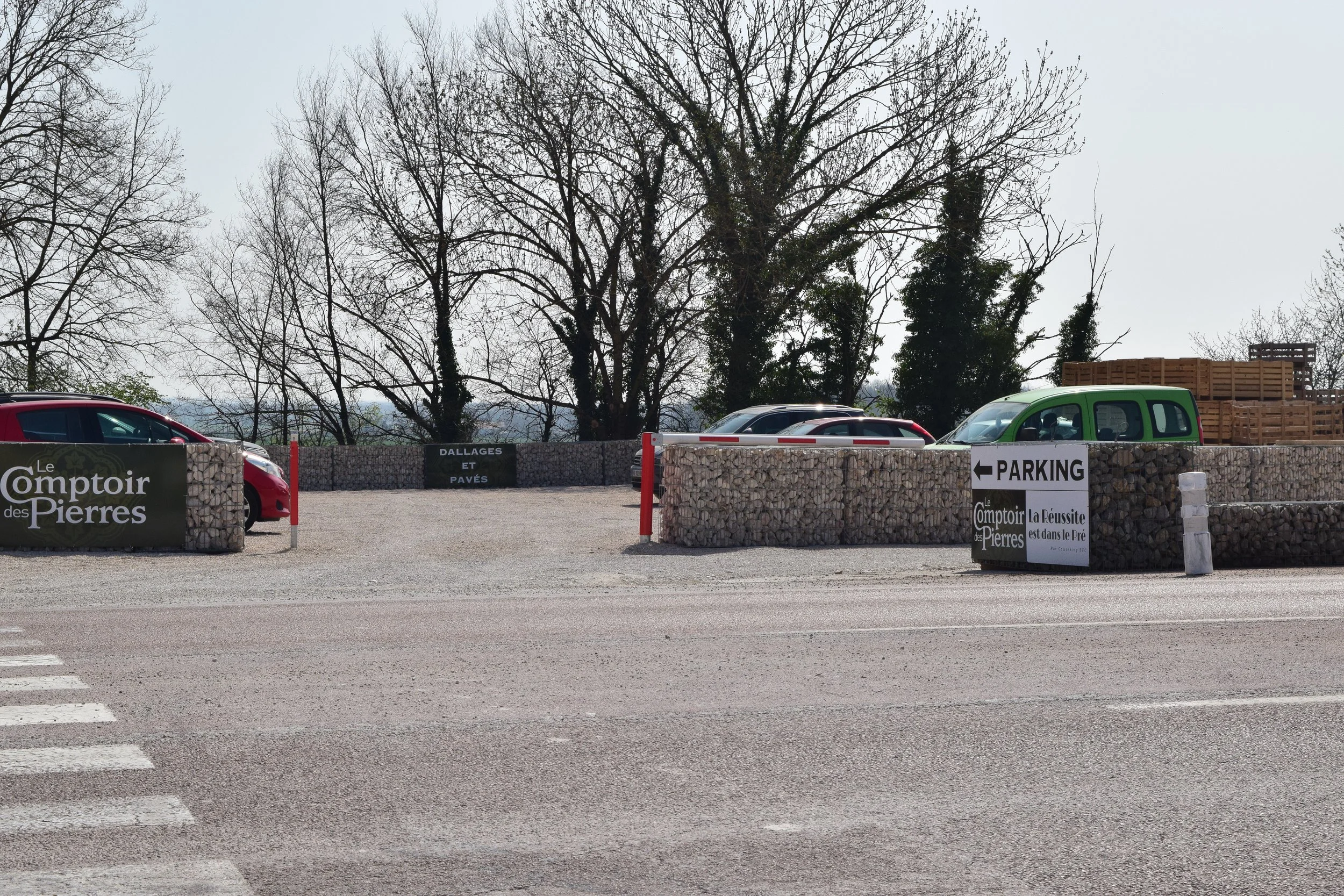 Un parking avec plusieurs voitures, un mur en gabions avec des panneaux publicitaires, et un fond avec des arbres sans feuilles.