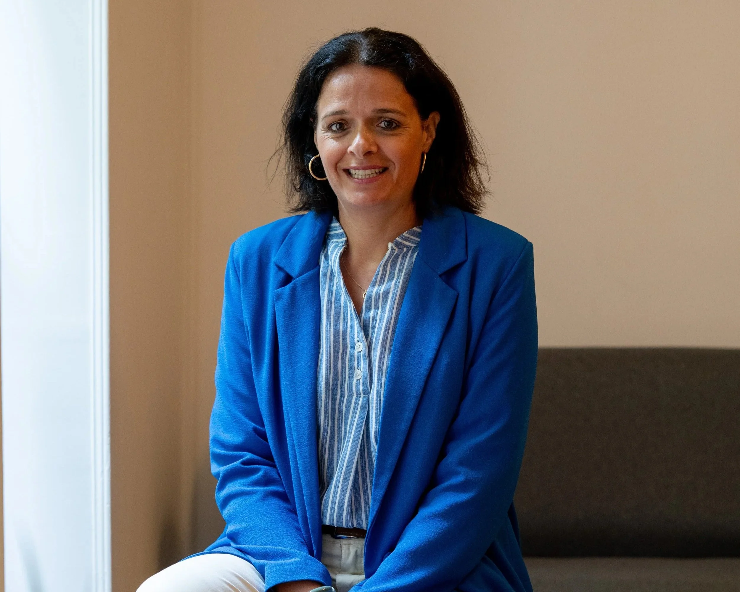 Une femme souriante avec des cheveux noirs porte un blazer bleu et une chemise à rayures. Elle est assise dans un intérieur avec un mur beige en arrière-plan.