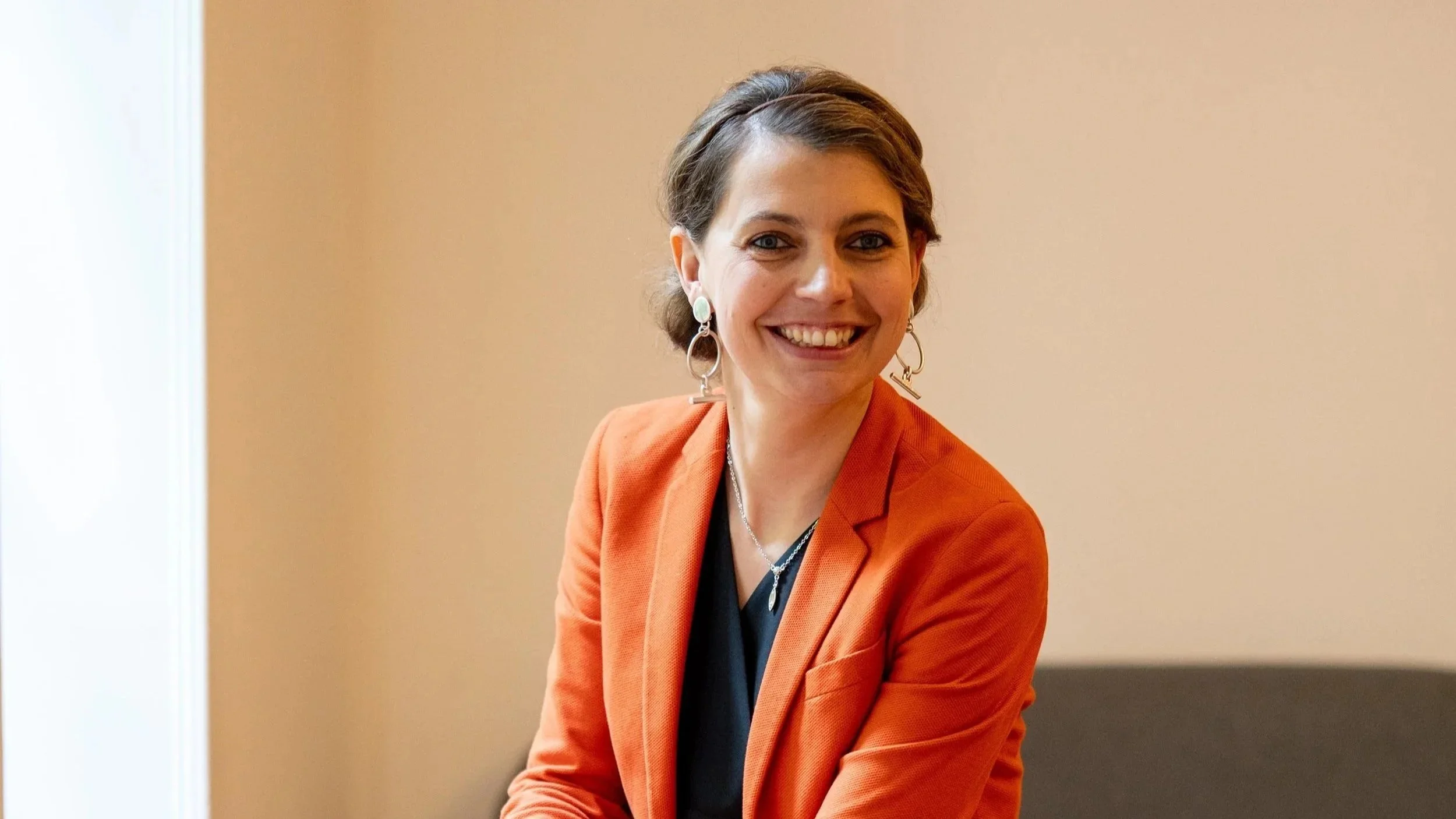 Une femme souriante avec des cheveux courts bruns, portant un blazer orange et des boucles d'oreilles pendantes, assise dans une pièce avec un mur beige en arrière-plan.