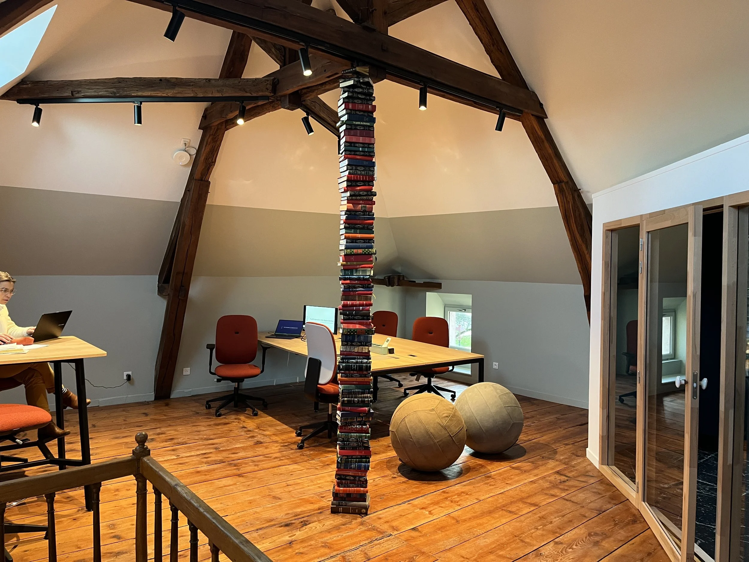 Une salle de réunion avec un grand tableau rond, des fauteuils orange, des ballons, et une structure en forme de tour faite de livres empilés, dans une pièce avec poutres en bois et fenêtres.