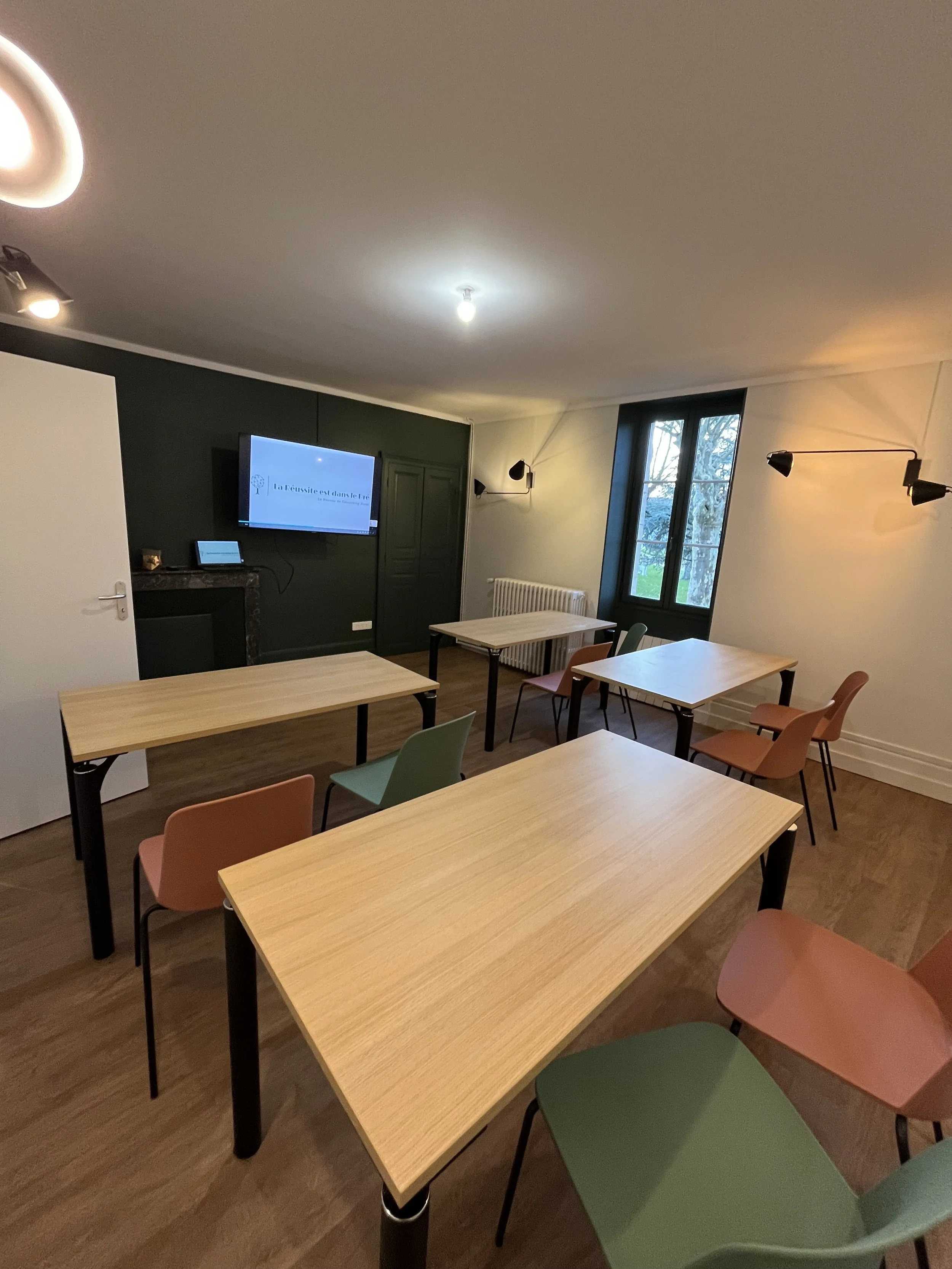 Une salle de réunion avec plusieurs tables en bois, des chaises de différentes couleurs, une télévision accrochée au mur avec un message écrit en français, deux lampes murales et une fenêtre donnant sur l'extérieur.